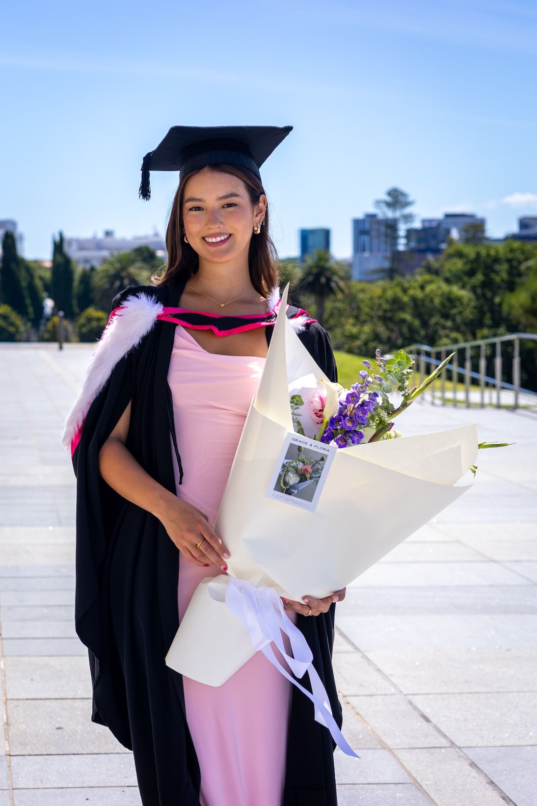 Smiling graduate in black cap and gown holding flower bouquet outdoors on a sunny day.
