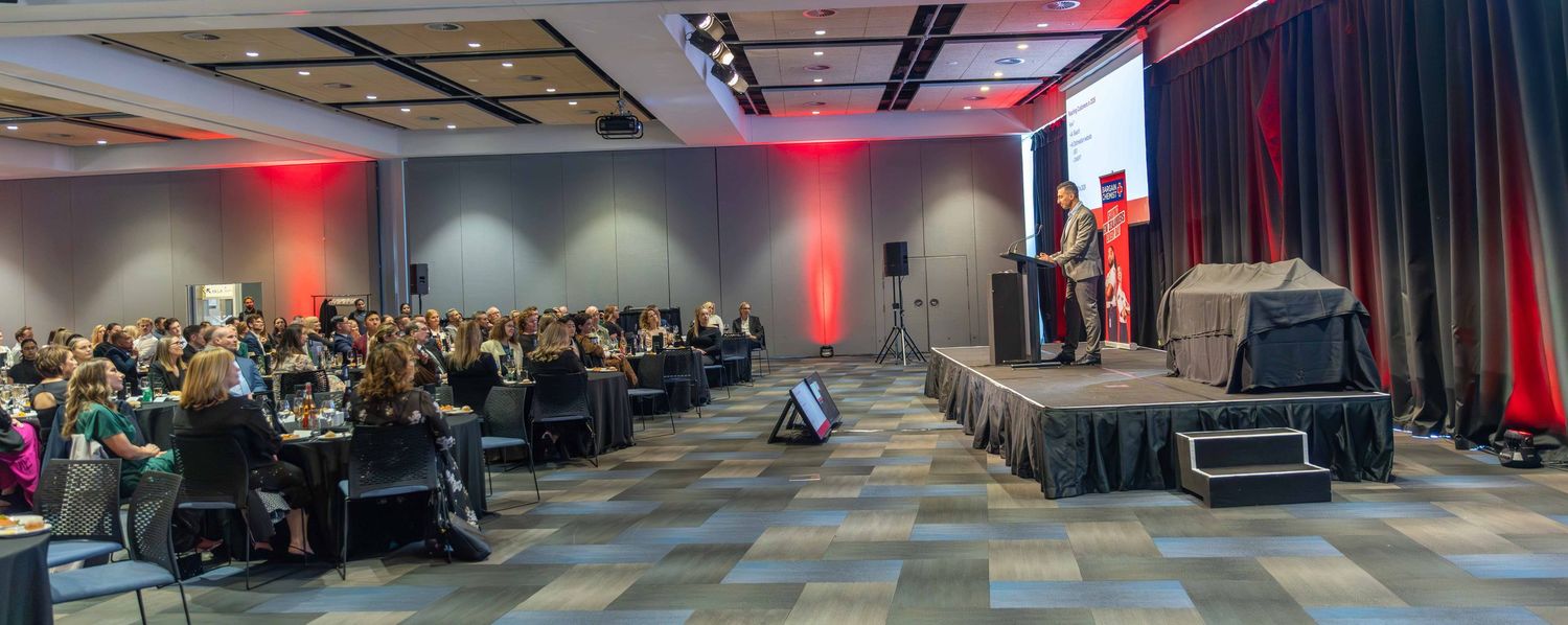Wide view of a corporate conference with attendees seated facing a stage with red accent lighting.