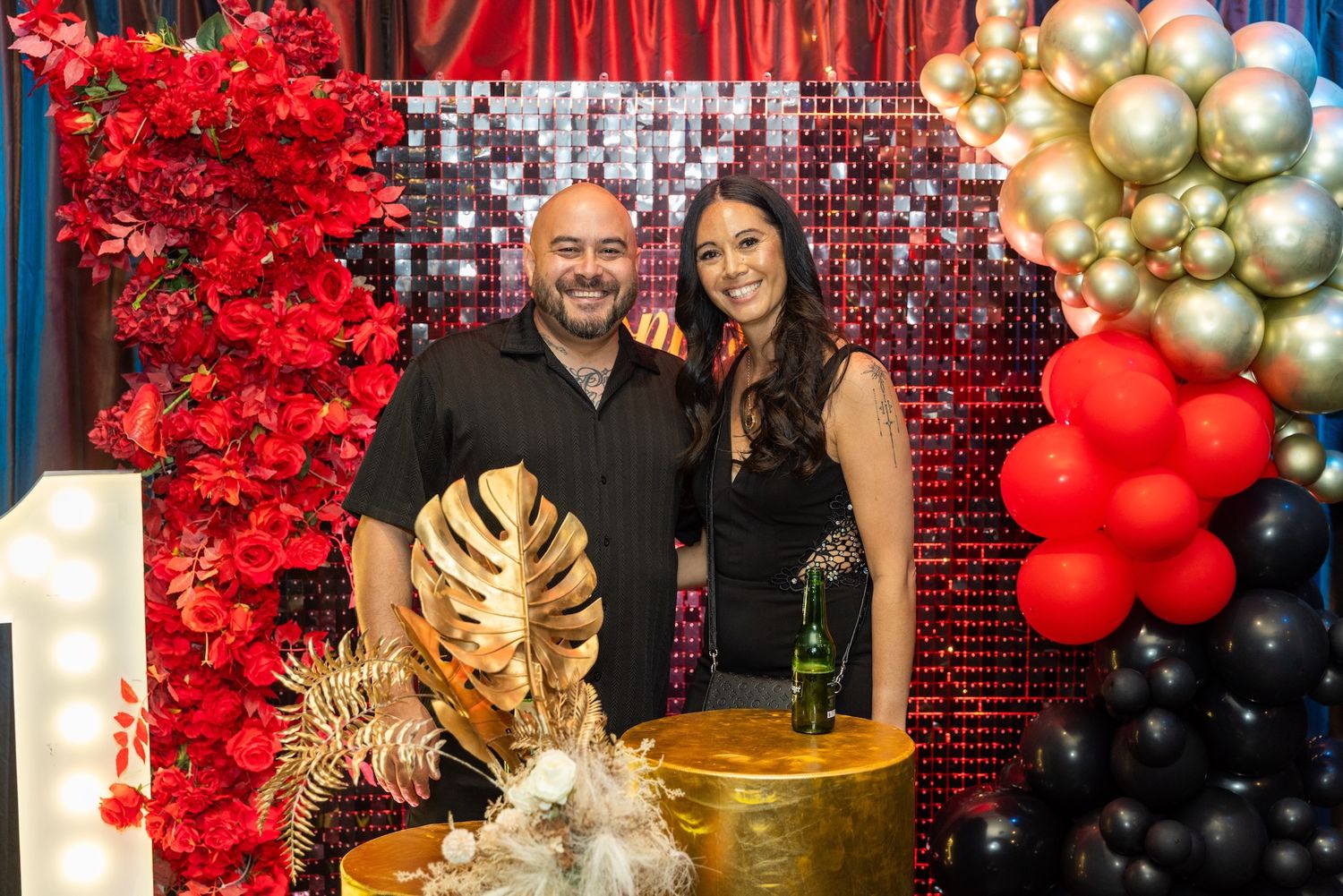 Couple posing at a festive party backdrop with red flowers, gold balloons, and a decorated cake table.