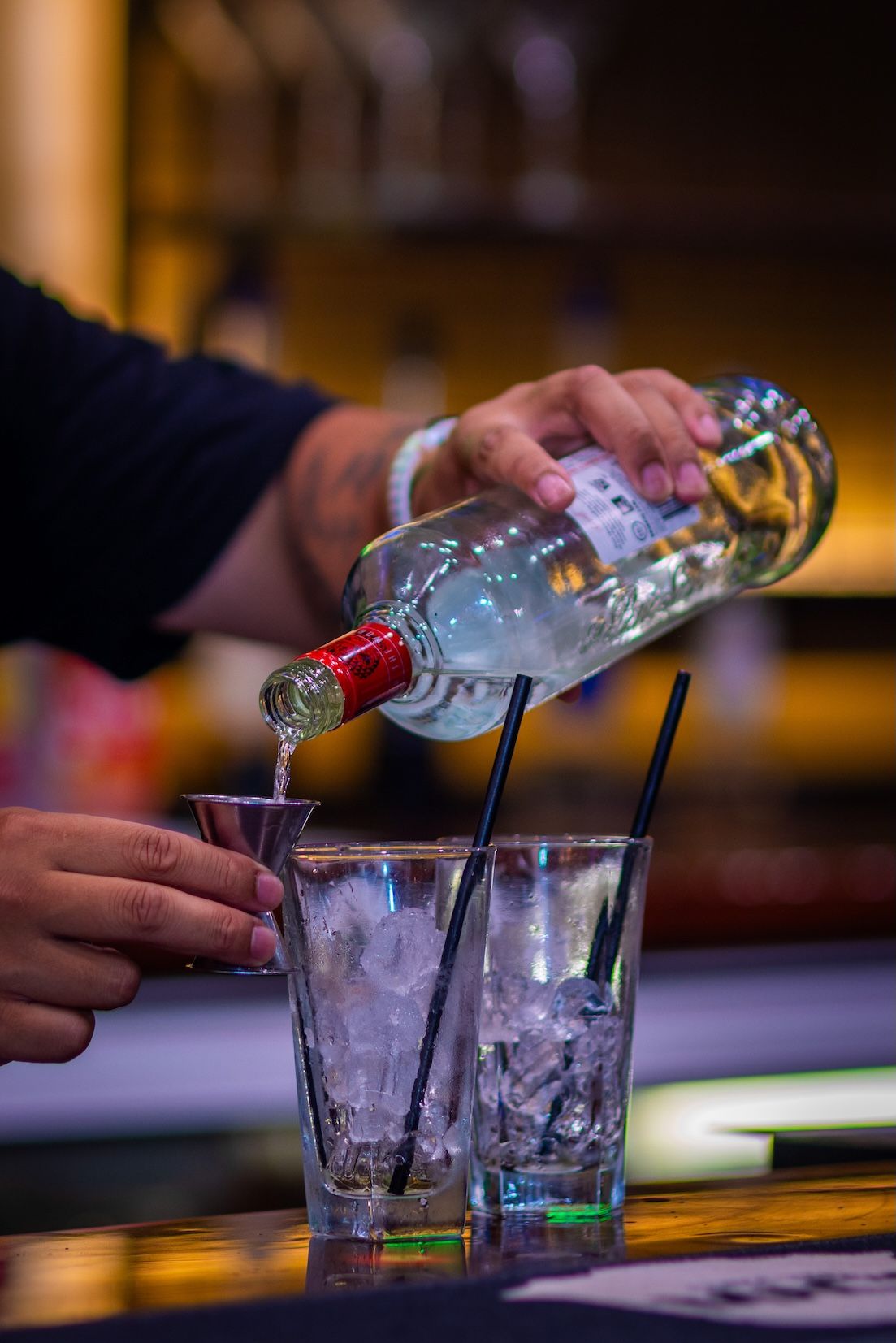 Clear liquor being poured into multiple glass cups with ice at a dimly lit bar.