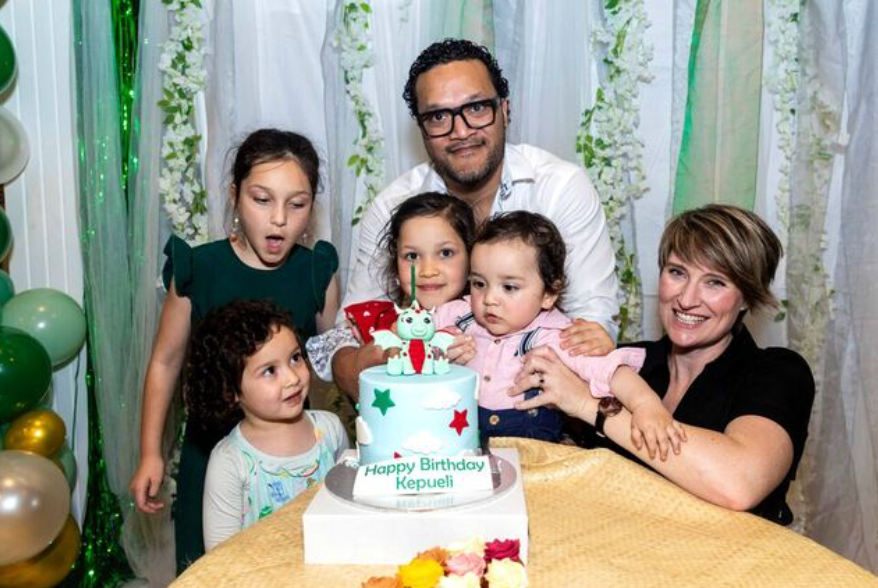 Family celebrating a birthday with a decorated cake reading Happy Birthday at a festive party with green and white balloons.