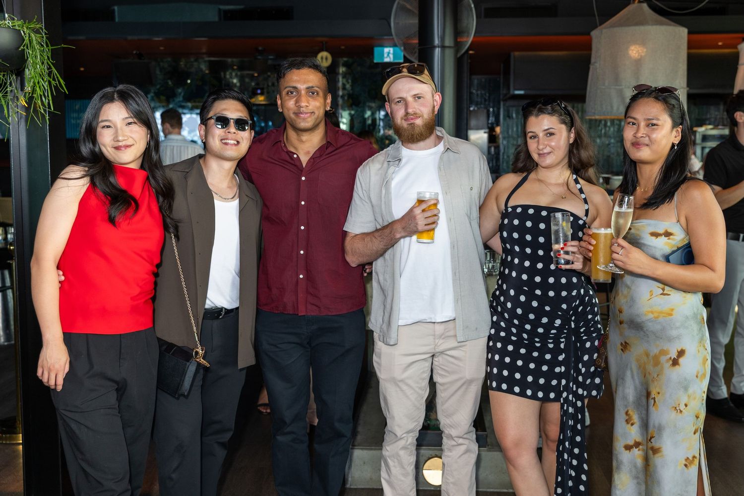Group of six friends smiling and holding drinks together at a casual indoor social gathering.