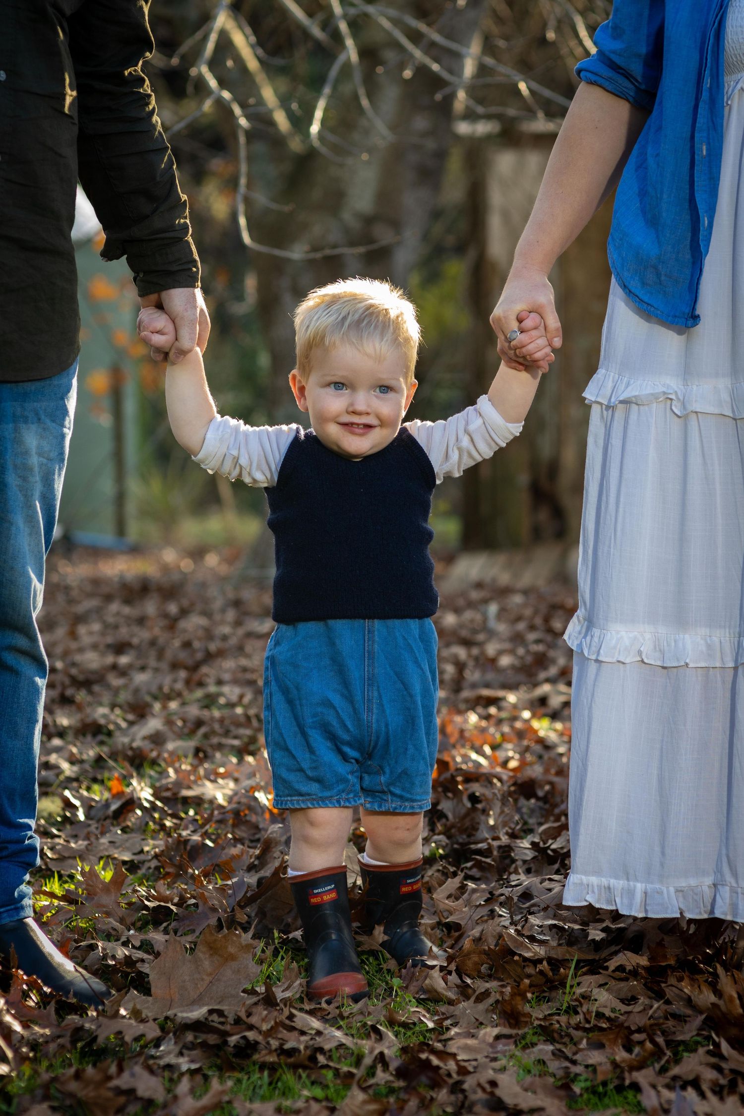 Smiling toddler boy holding parents hands standing on autumn leaves in a wooded outdoor setting.