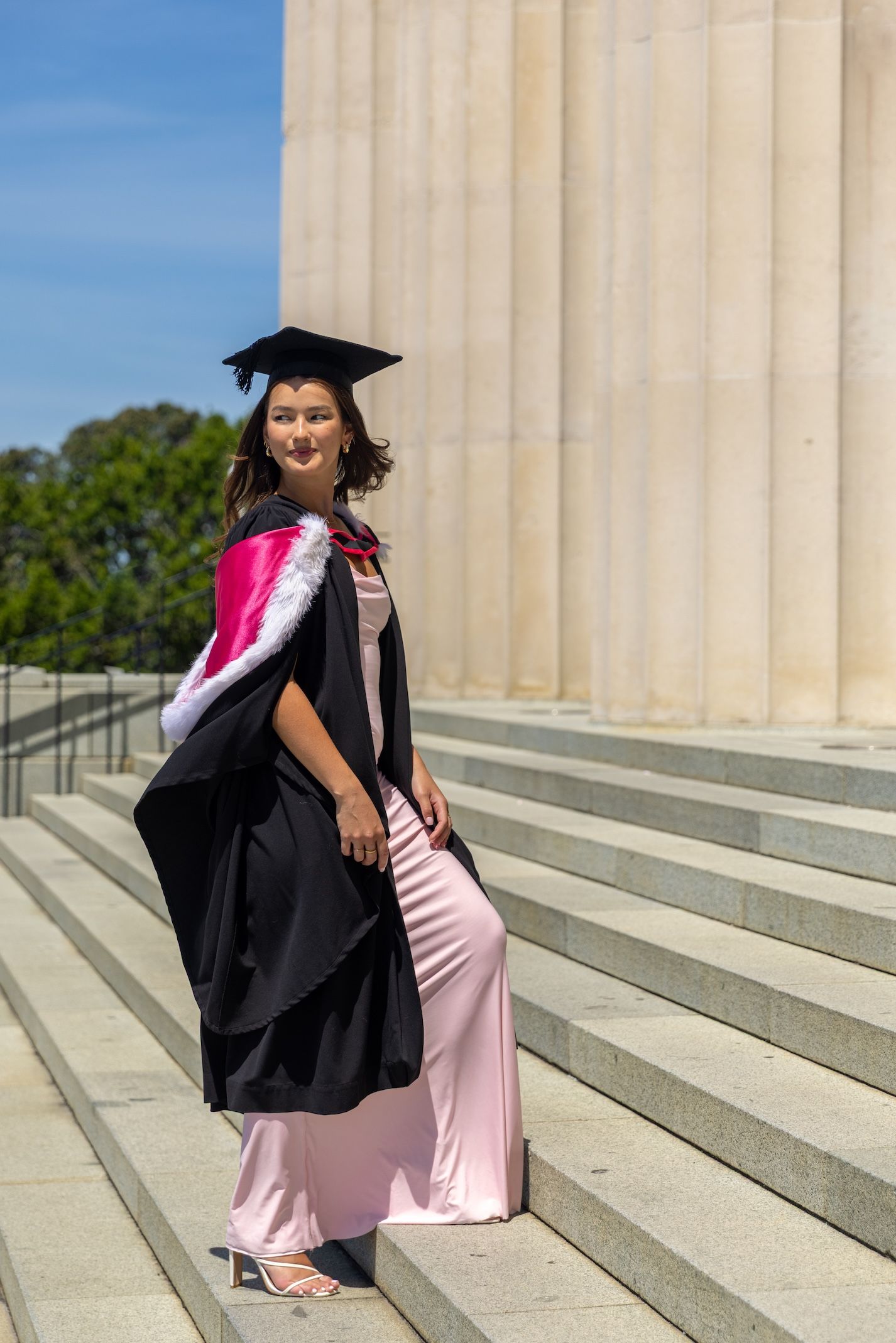 Young woman in black graduation gown and cap poses on stone steps beside large columns on a sunny day.