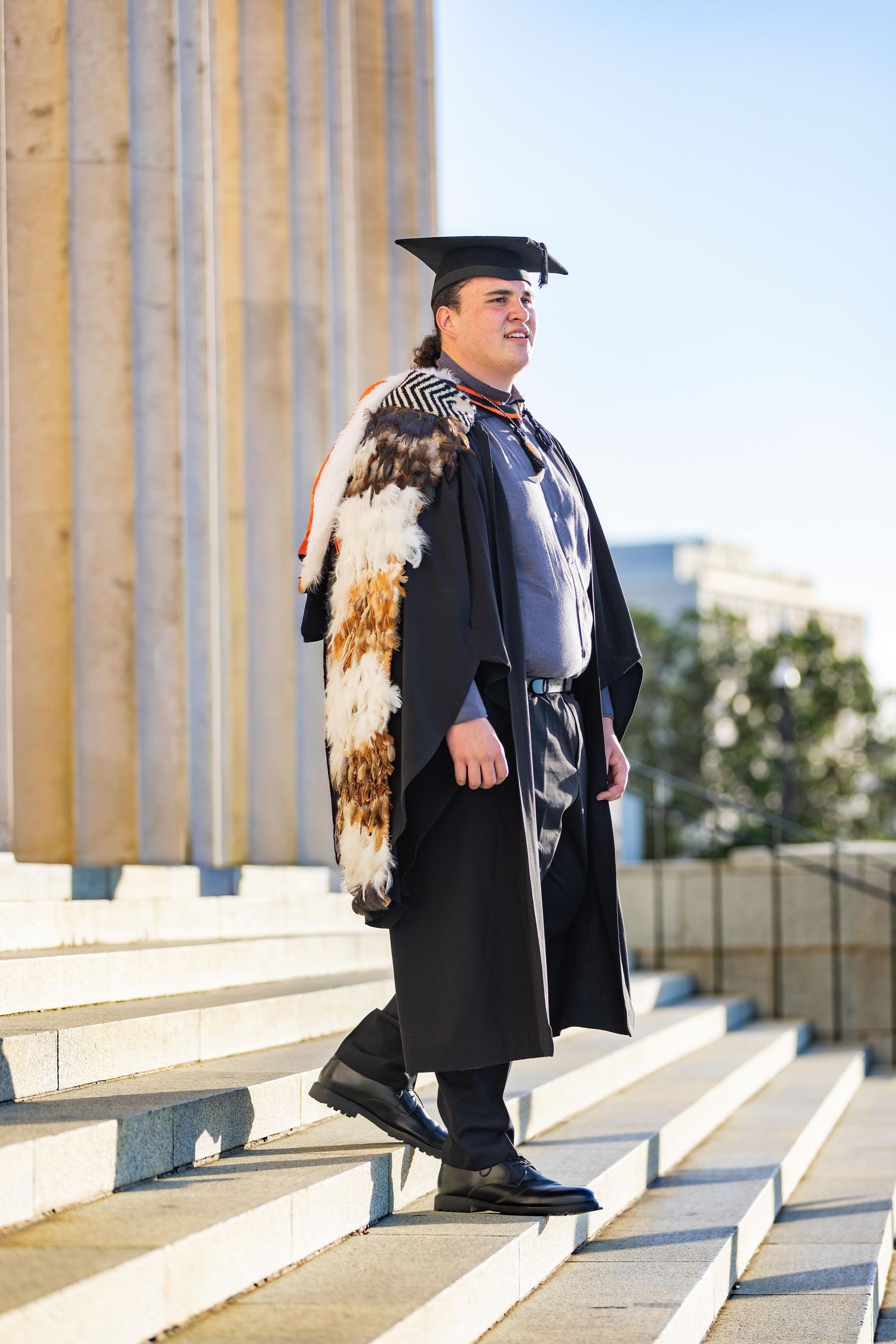 Graduate in cap and gown with traditional Maori korowai cloak stands proudly on university steps.