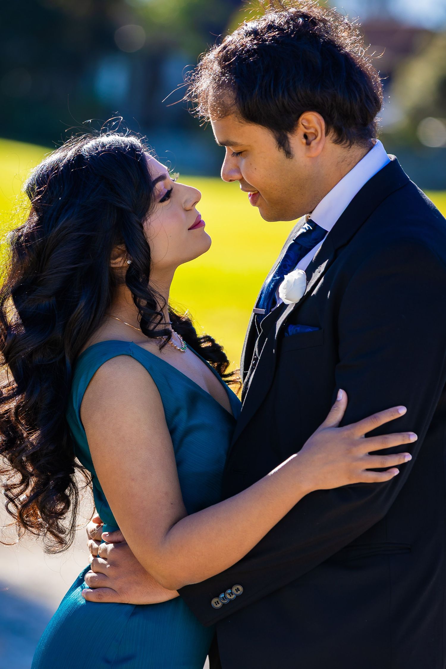 Couple in formal attire embracing outdoors, woman in teal dress and man in dark suit with boutonniere, sharing a romantic moment.