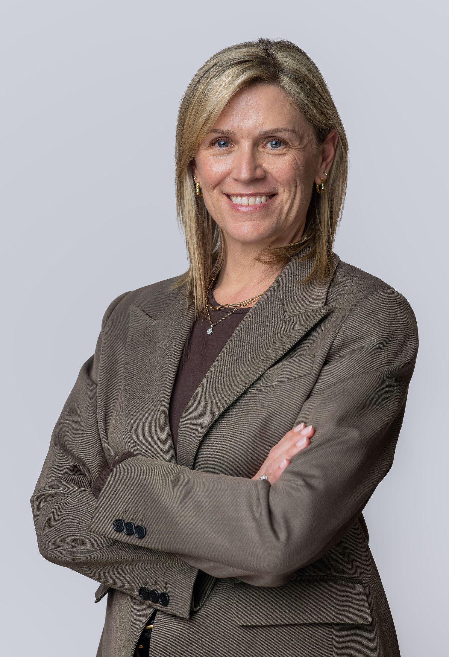 Professional woman in tan blazer with arms crossed, smiling confidently against gray background.