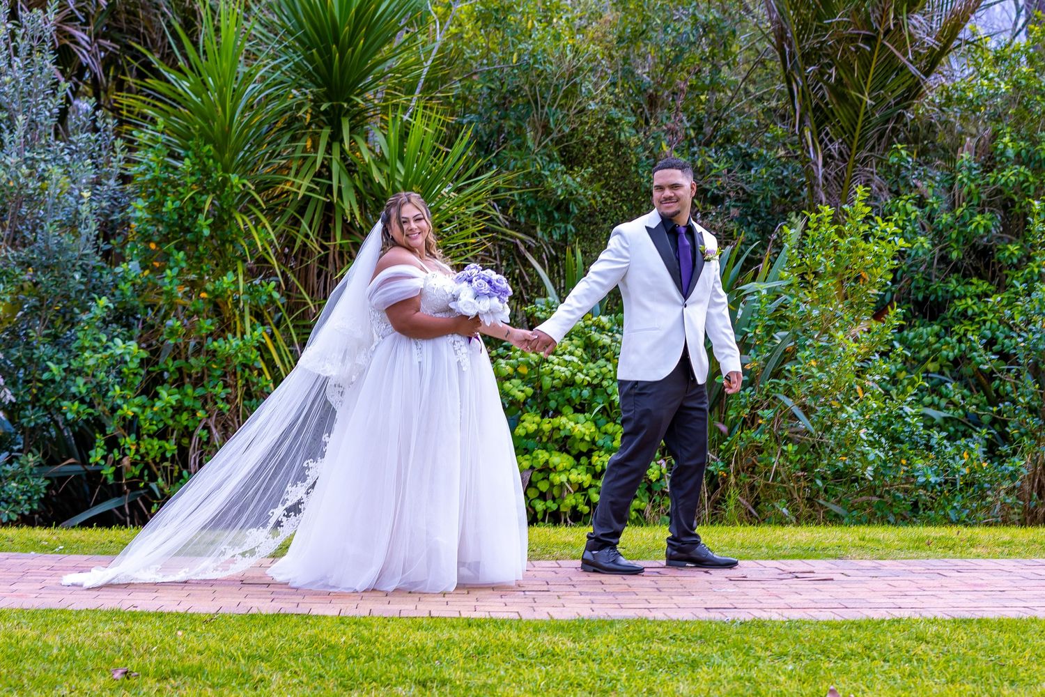 Bride and groom holding hands on brick path in lush garden, bride in white gown with veil, groom in white blazer.