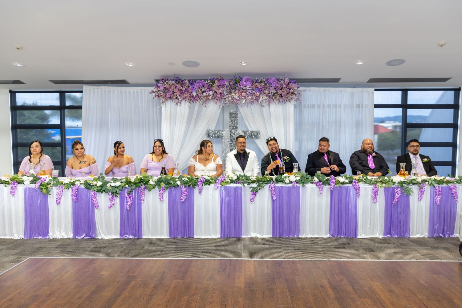 Wedding party seated at a decorated head table with purple and white floral arrangements in an elegant reception venue.