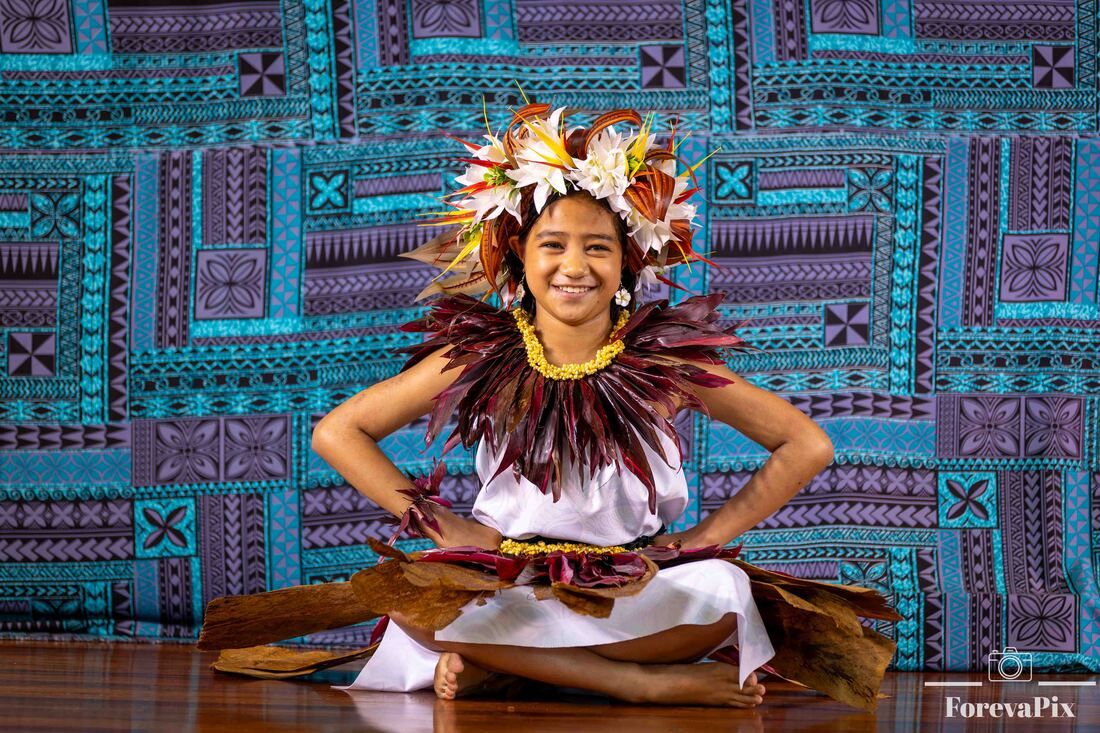 Young Polynesian dancer in traditional costume with floral headdress and feather lei performs seated pose.