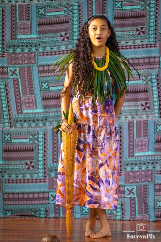 Young Polynesian girl in colorful floral dress with yellow lei and palm leaf adornments performs traditional dance on stage.