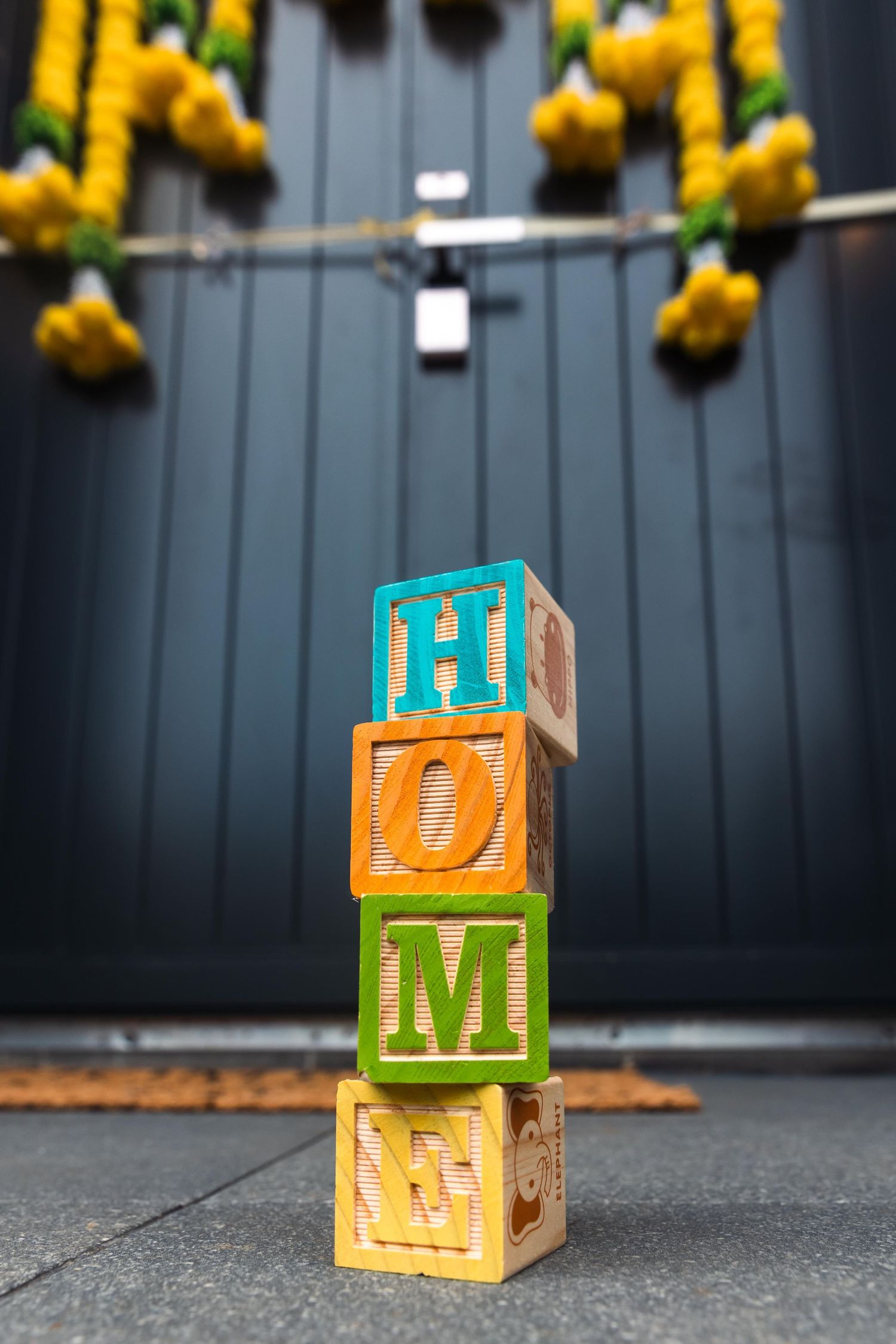 Colorful wooden alphabet blocks stacked to spell HOME on a doorstep in front of a dark door with yellow flowers.