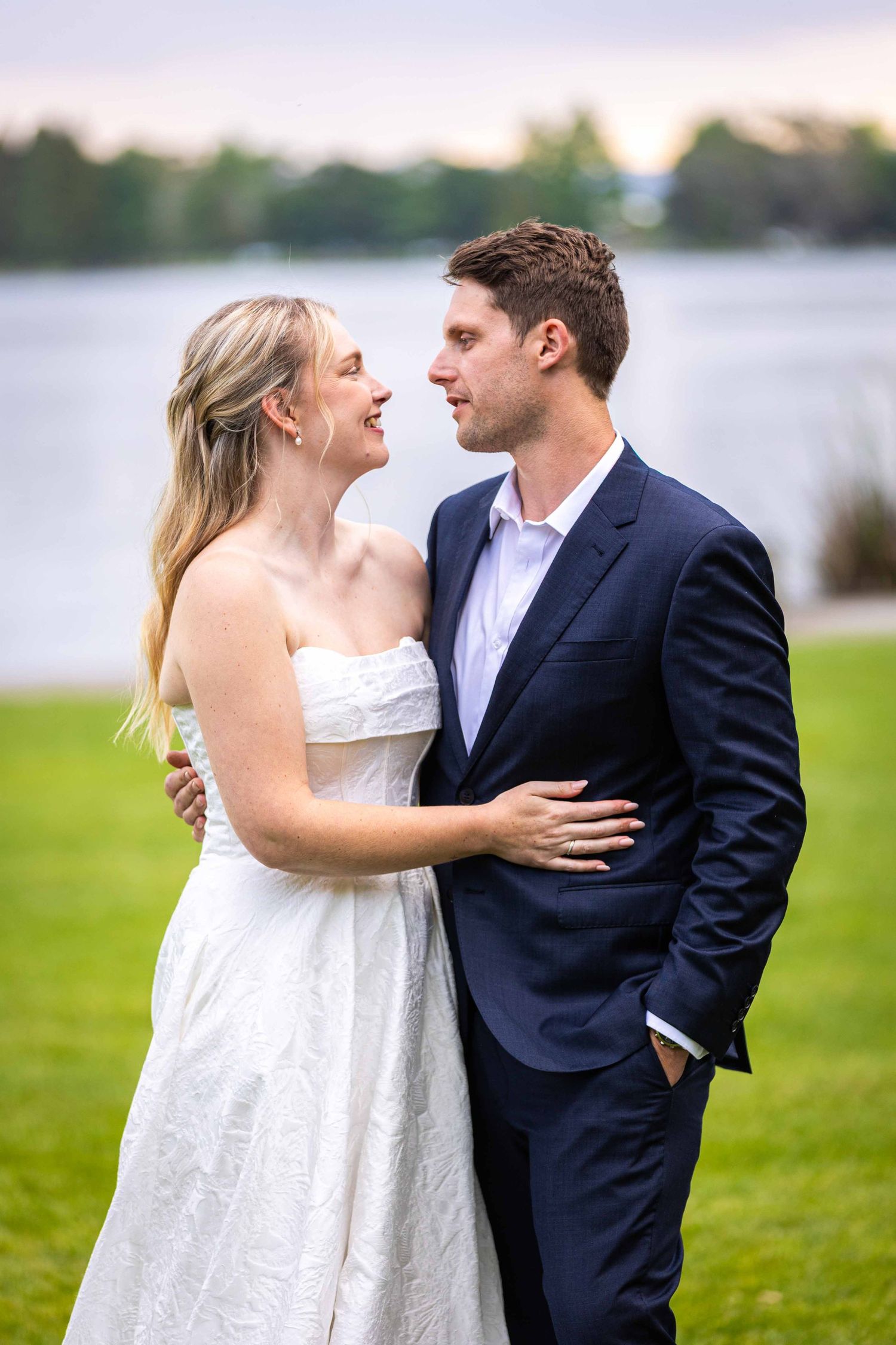A couple embraces on a grassy lawn with a lake in the background.