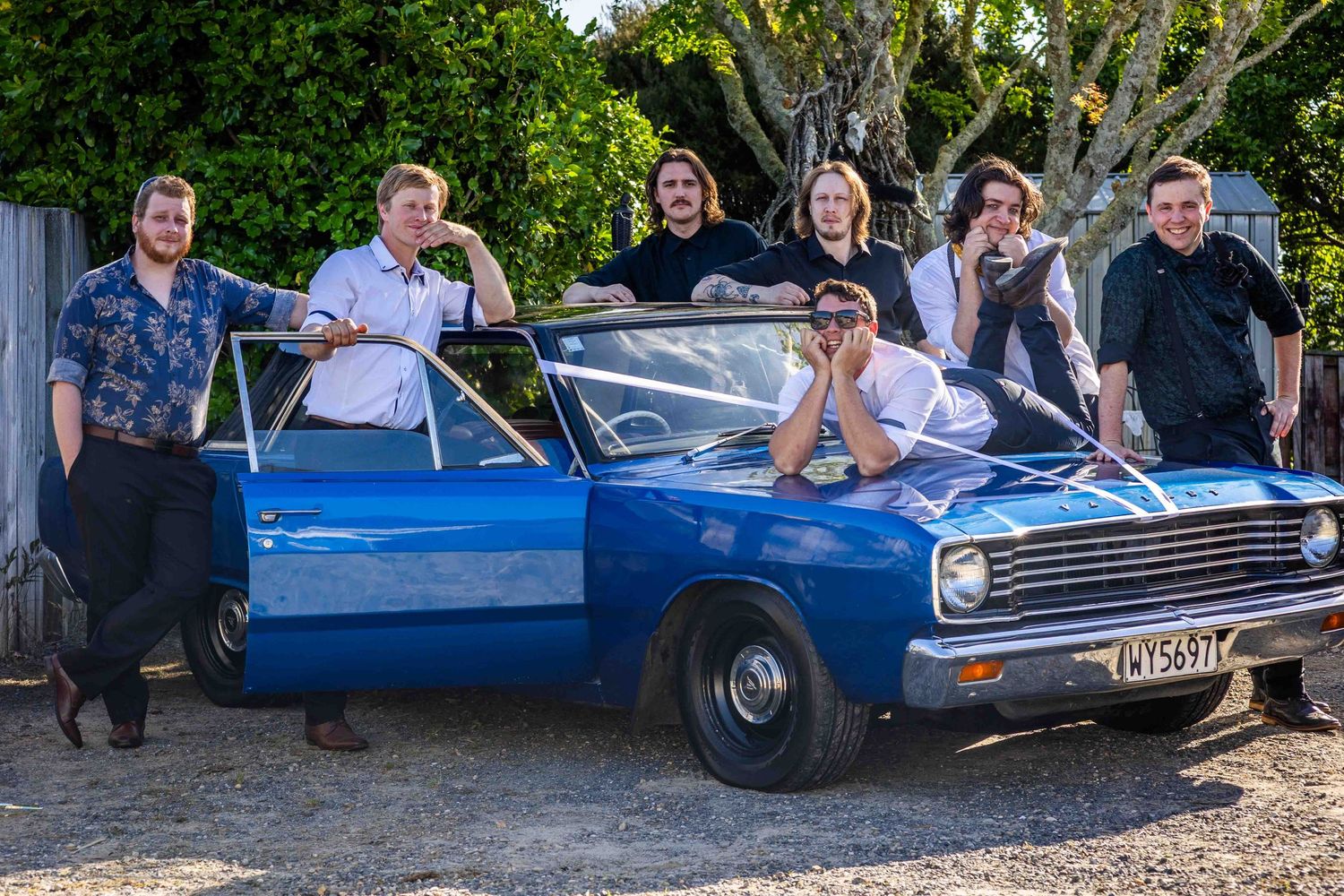 Seven men posing with a classic blue muscle car outdoors on a sunny day.