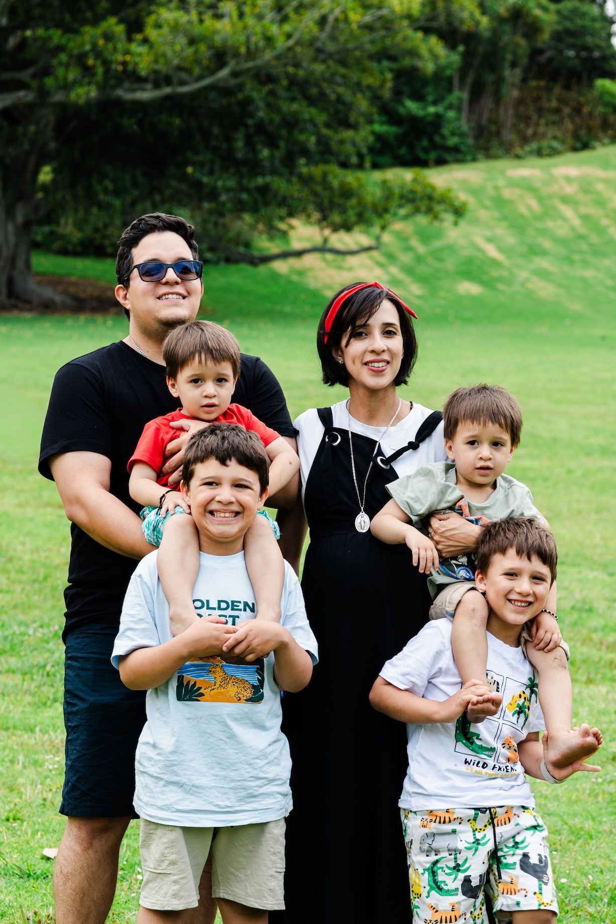 Family of six smiling outdoors on green grass, parents holding toddlers with two boys in front, trees in background.