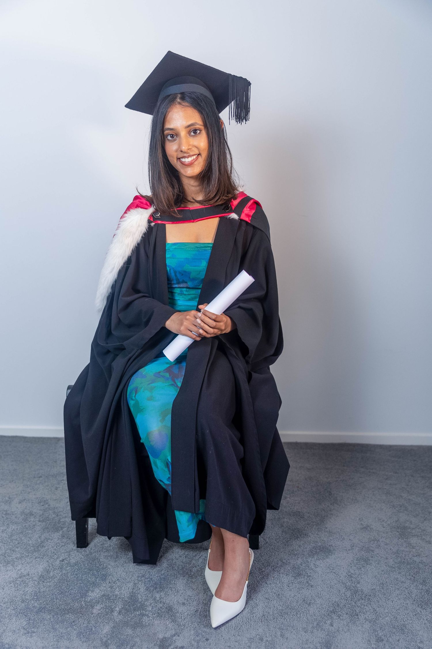 Smiling female graduate in black cap and gown holding diploma scroll, seated against light gray background.