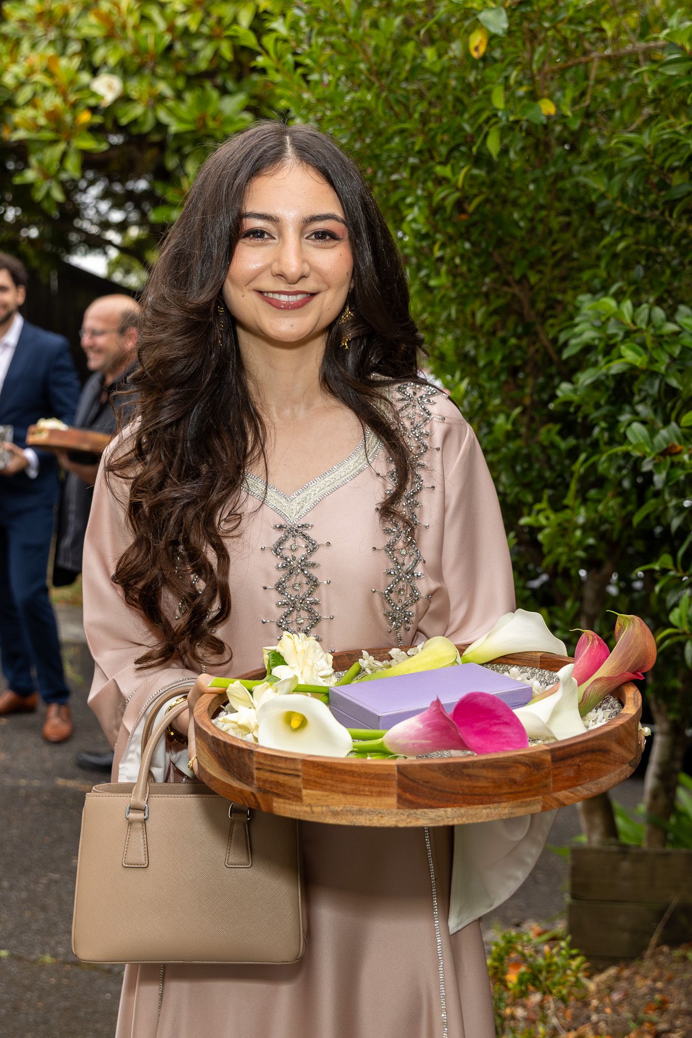 Young woman in beige embroidered dress holding wooden tray of colorful flowers at outdoor event.