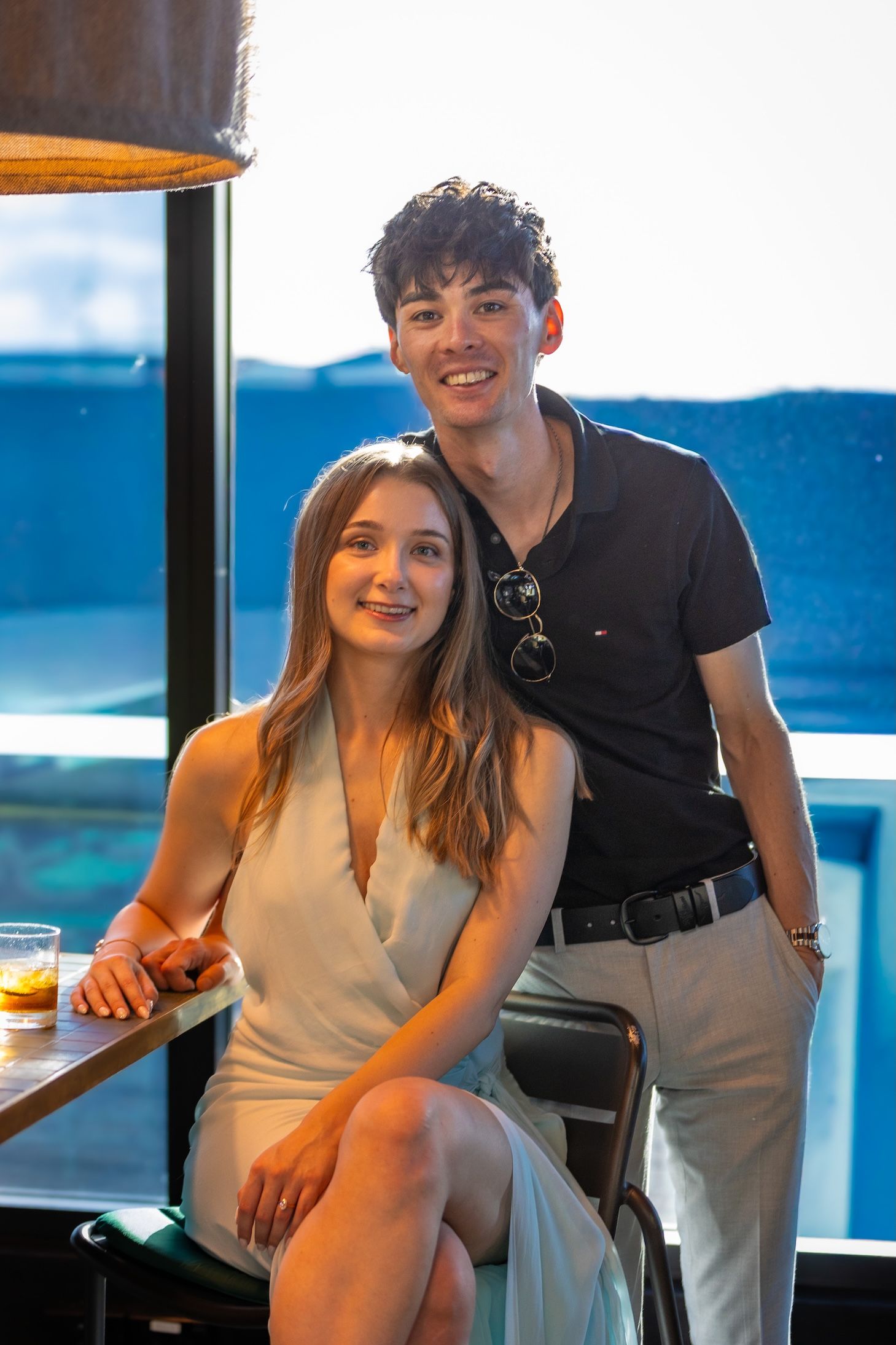 Young couple posing together at a rooftop bar, smiling, with a scenic blue view in the background.