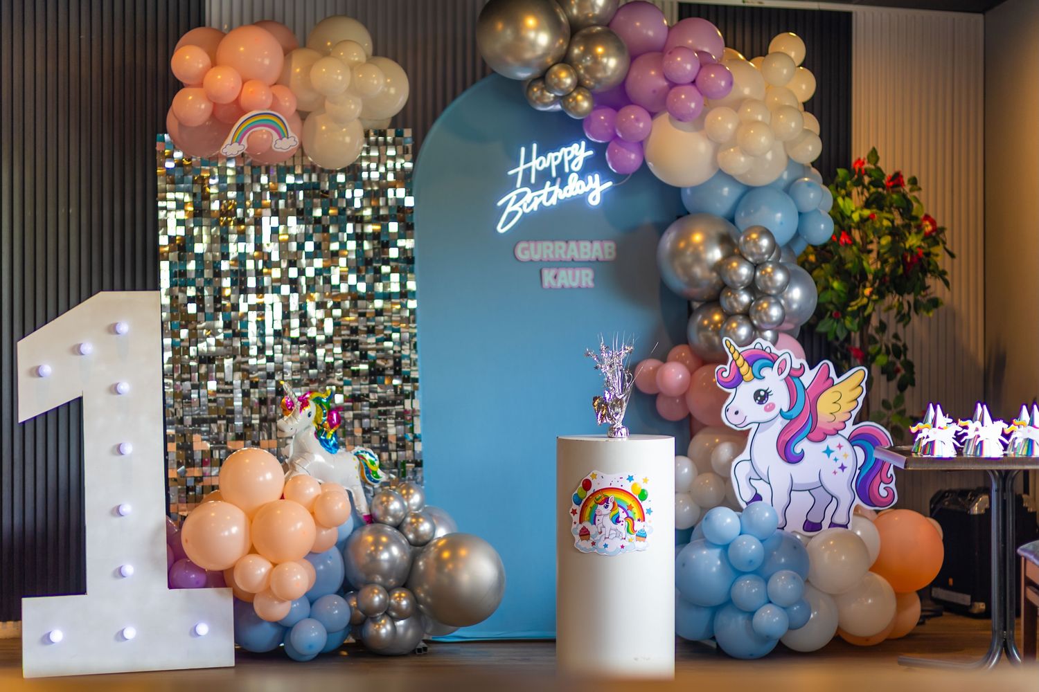Unicorn-themed first birthday party setup with balloon arch, neon happy birthday sign, and decorative number one light.