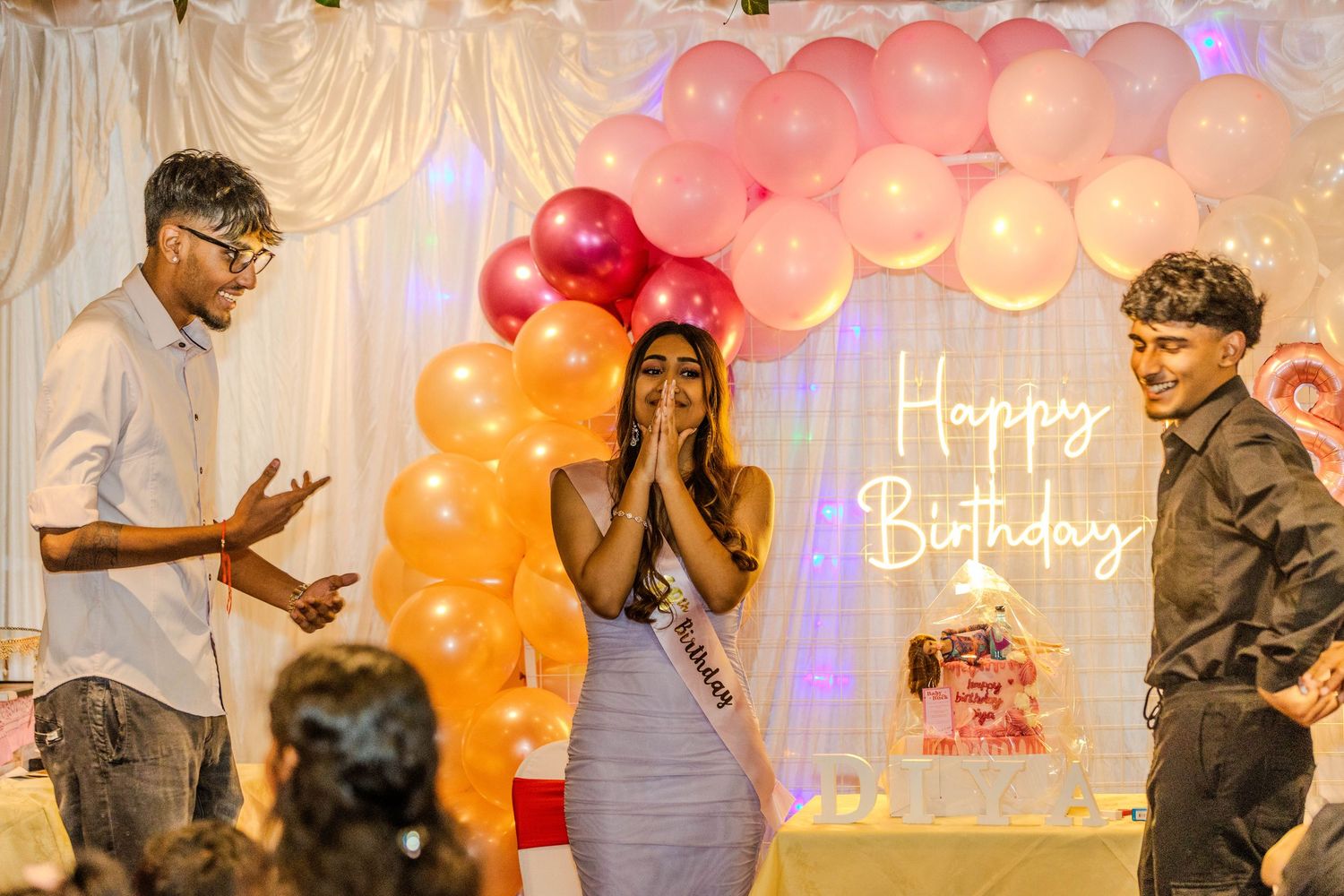 Woman surprised at birthday party with pink balloon arch and Happy Birthday sign, flanked by two men.