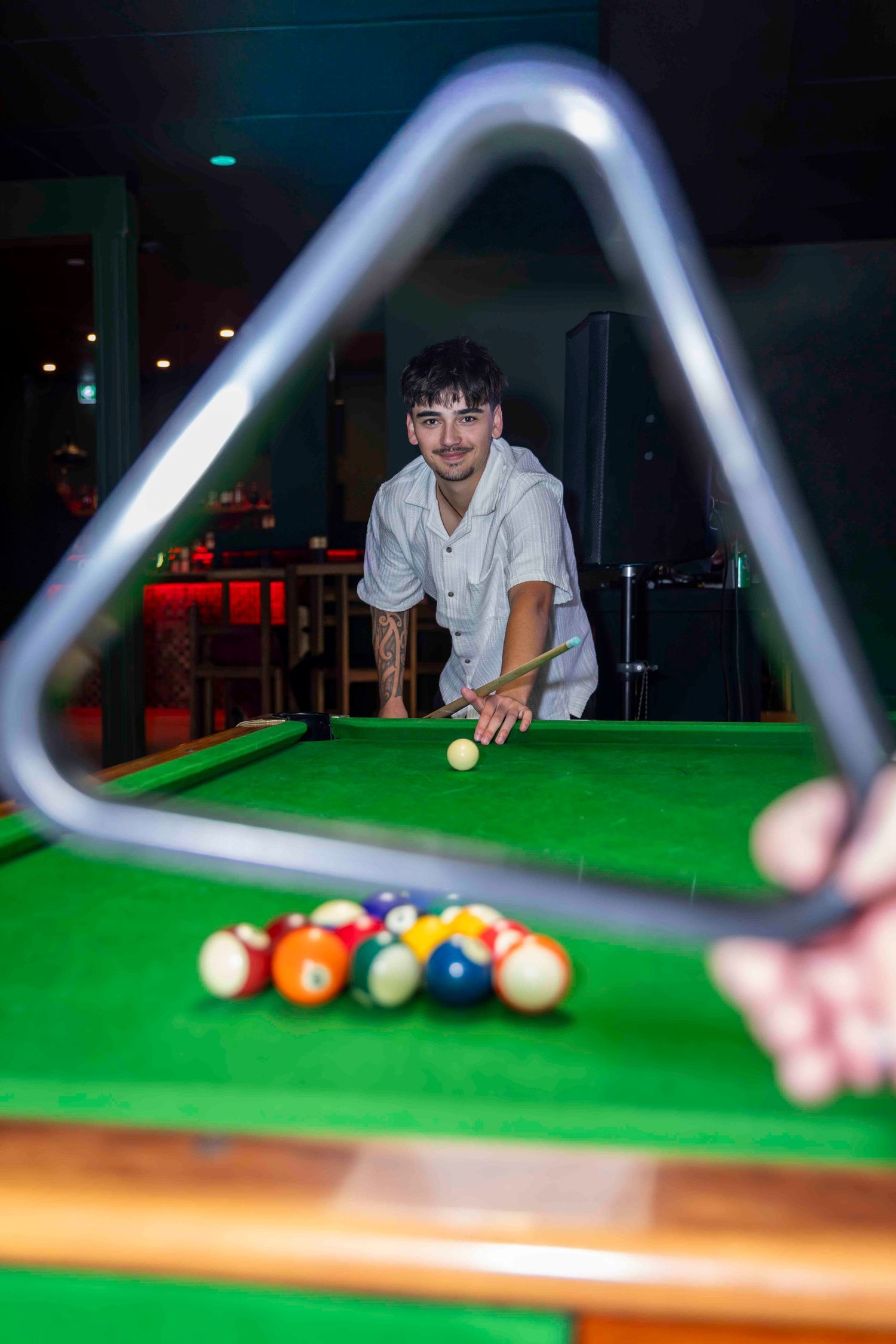 Young man with tattoos leans over green pool table holding cue stick, aiming at billiard balls through triangle rack.