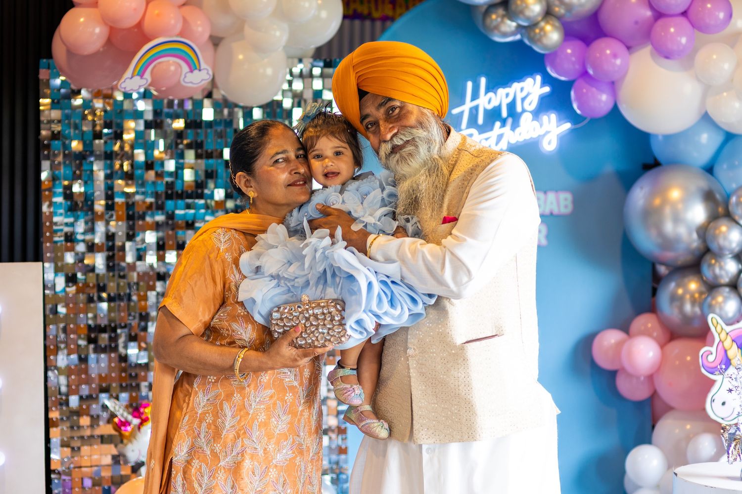 Sikh family celebrating a birthday, grandparents holding a baby in blue outfit, colorful balloon decorations backdrop.