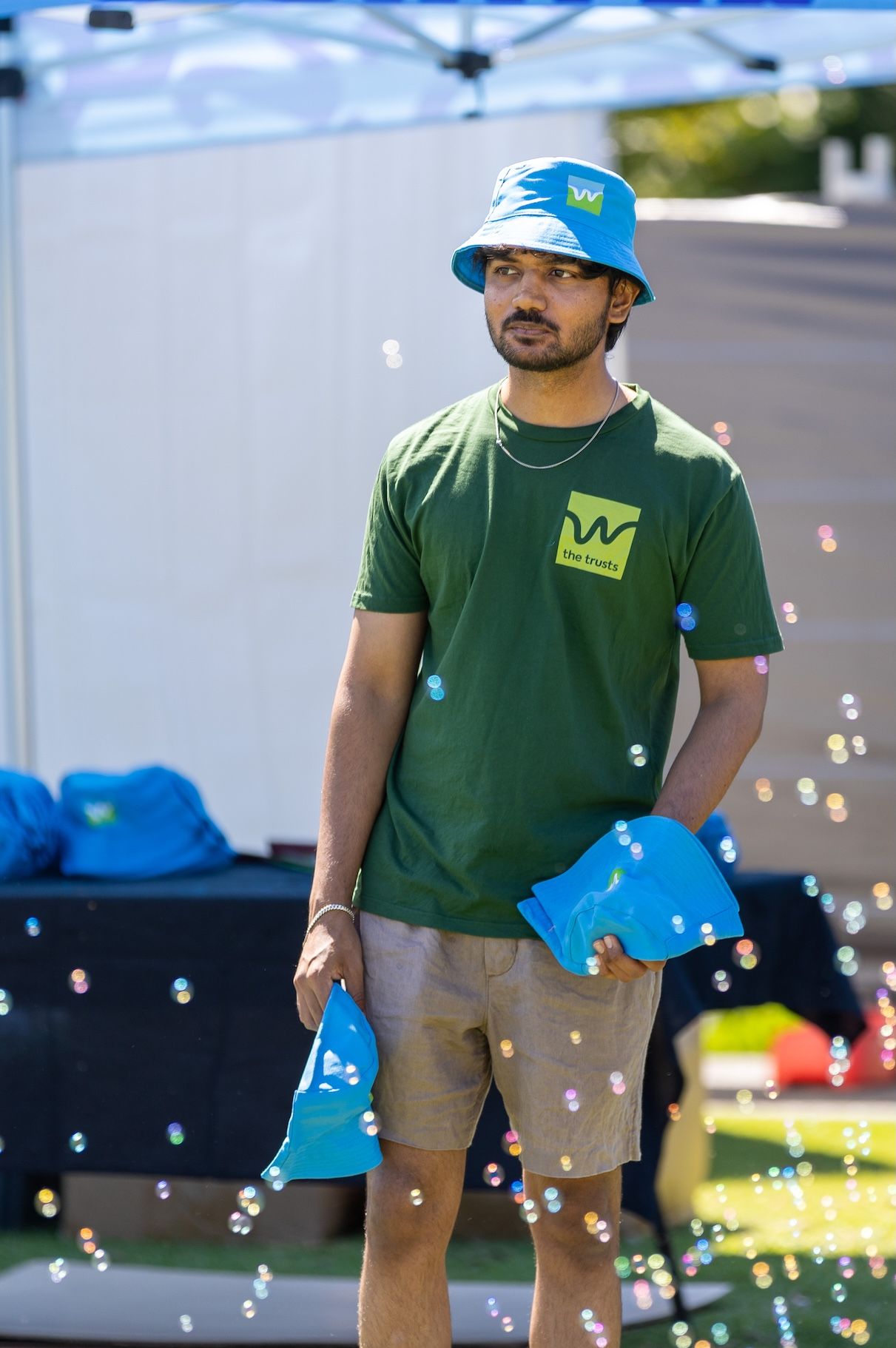 Staff member in green shirt and blue hat stands outdoors at event with sparkly bokeh effects.