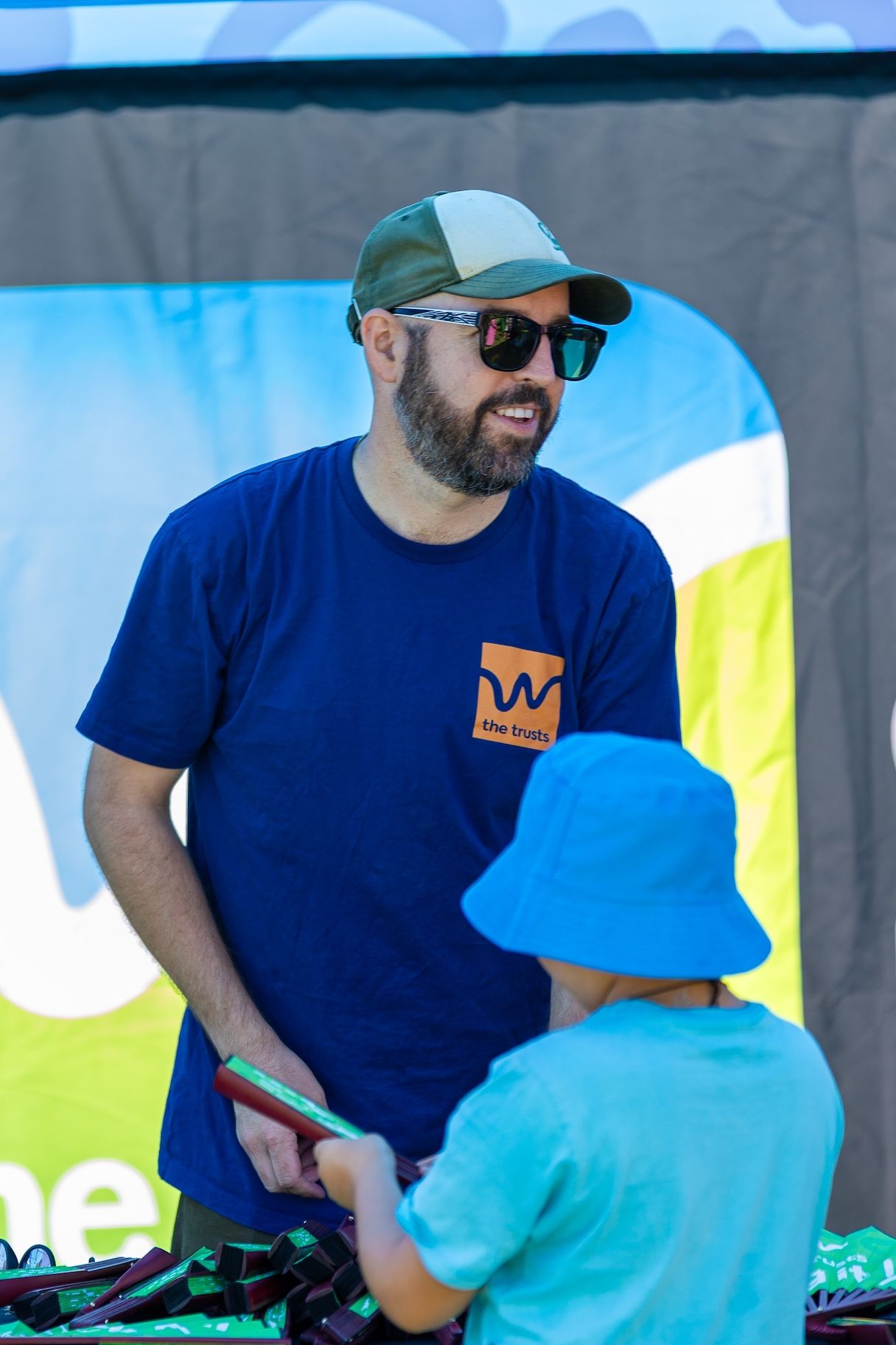 Person in navy blue t-shirt with crown logo interacting with child in bright blue cap at outdoor event.
