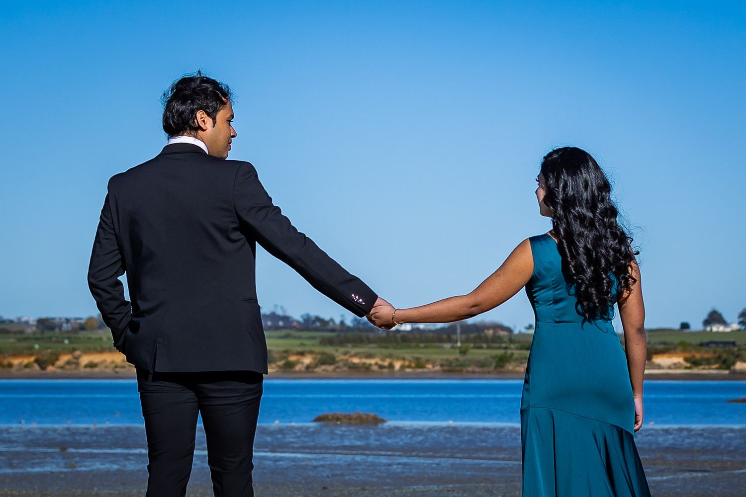 Couple in formal attire holding hands while gazing at a serene blue ocean under a clear sky.