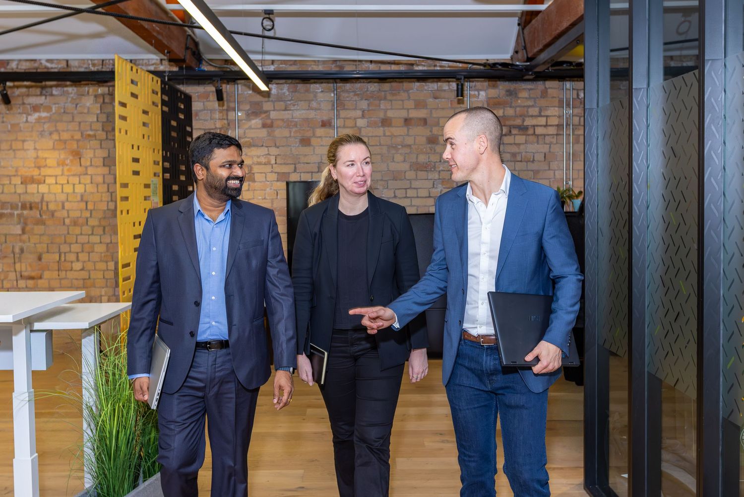 Three business professionals walking and talking in a modern brick-walled office space.