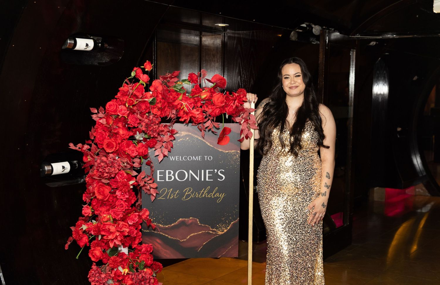 Woman in gold sequin gown poses beside floral arch and welcome sign at Ebonie's 21st birthday celebration.