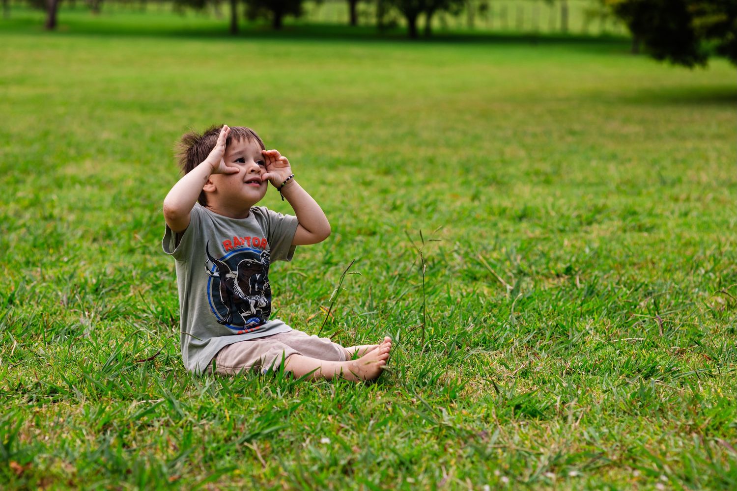 Young boy sitting cross-legged on lush green grass, shielding his eyes from the sun on a bright day.