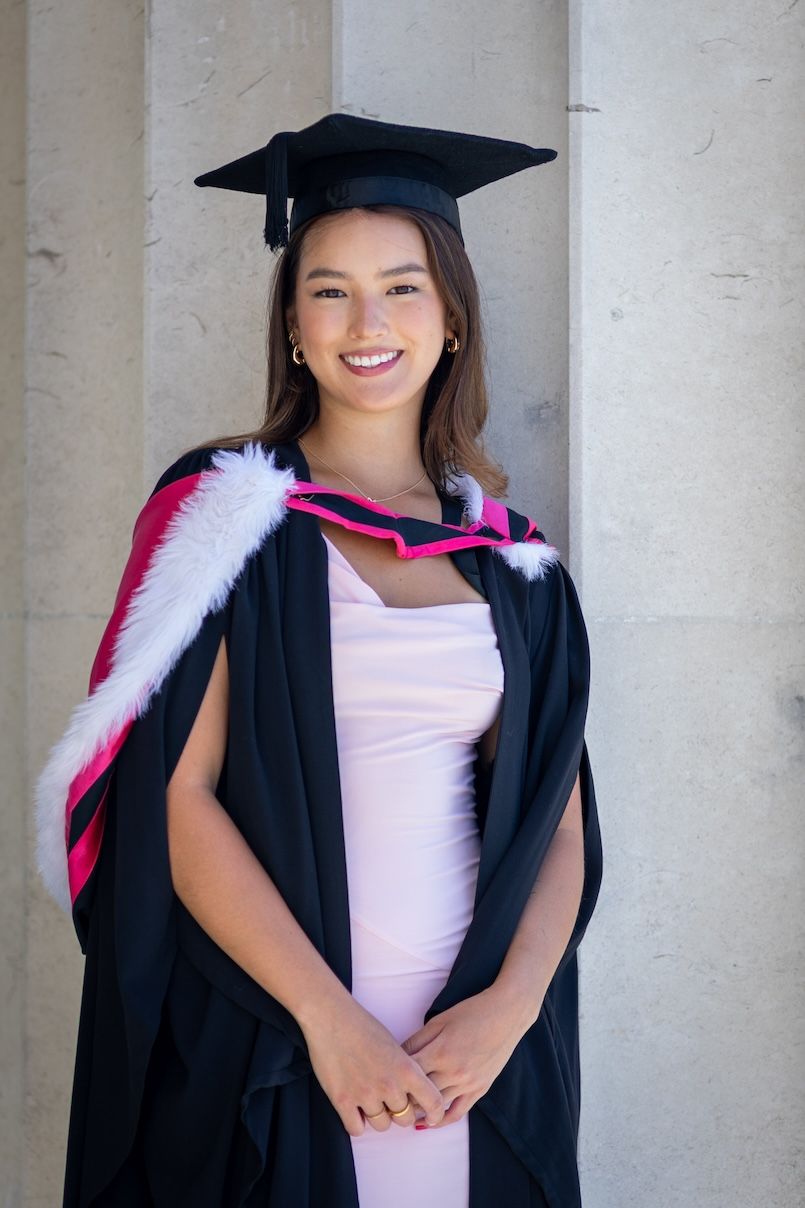Smiling young woman in black graduation gown with pink trim and mortarboard cap posing between stone columns.