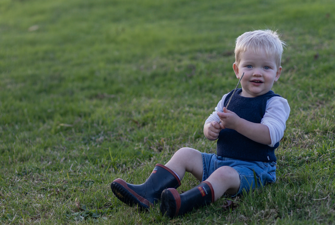 Smiling toddler boy sitting on green grass wearing navy vest, blue shorts, and rain boots.
