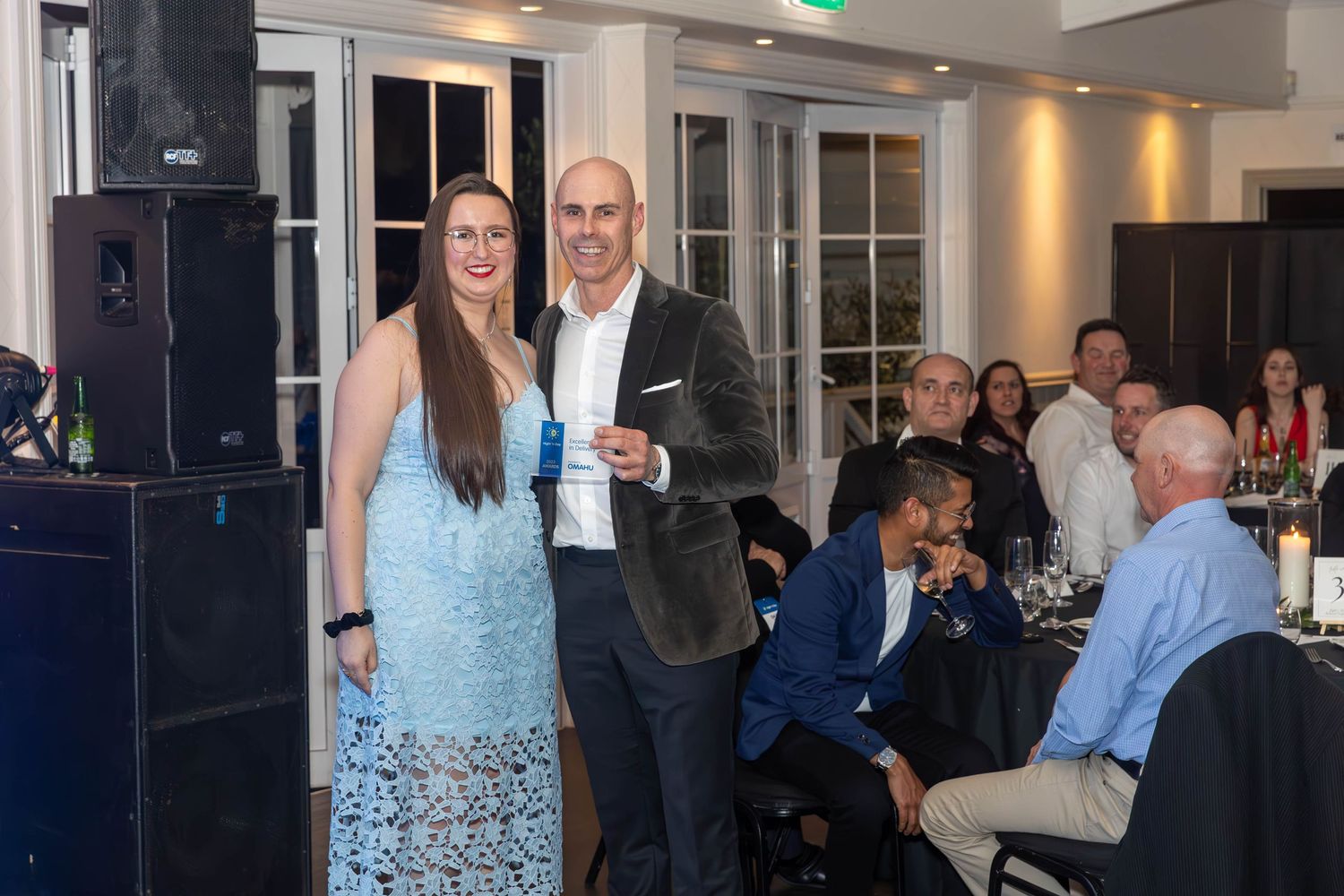 A man and woman in formal attire smile at a gala event with guests seated at tables in the background.