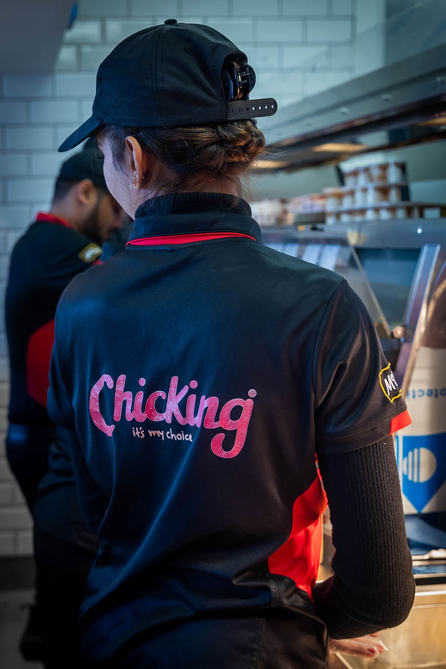 A Chicking restaurant employee in a black uniform with pink logo works in the kitchen.