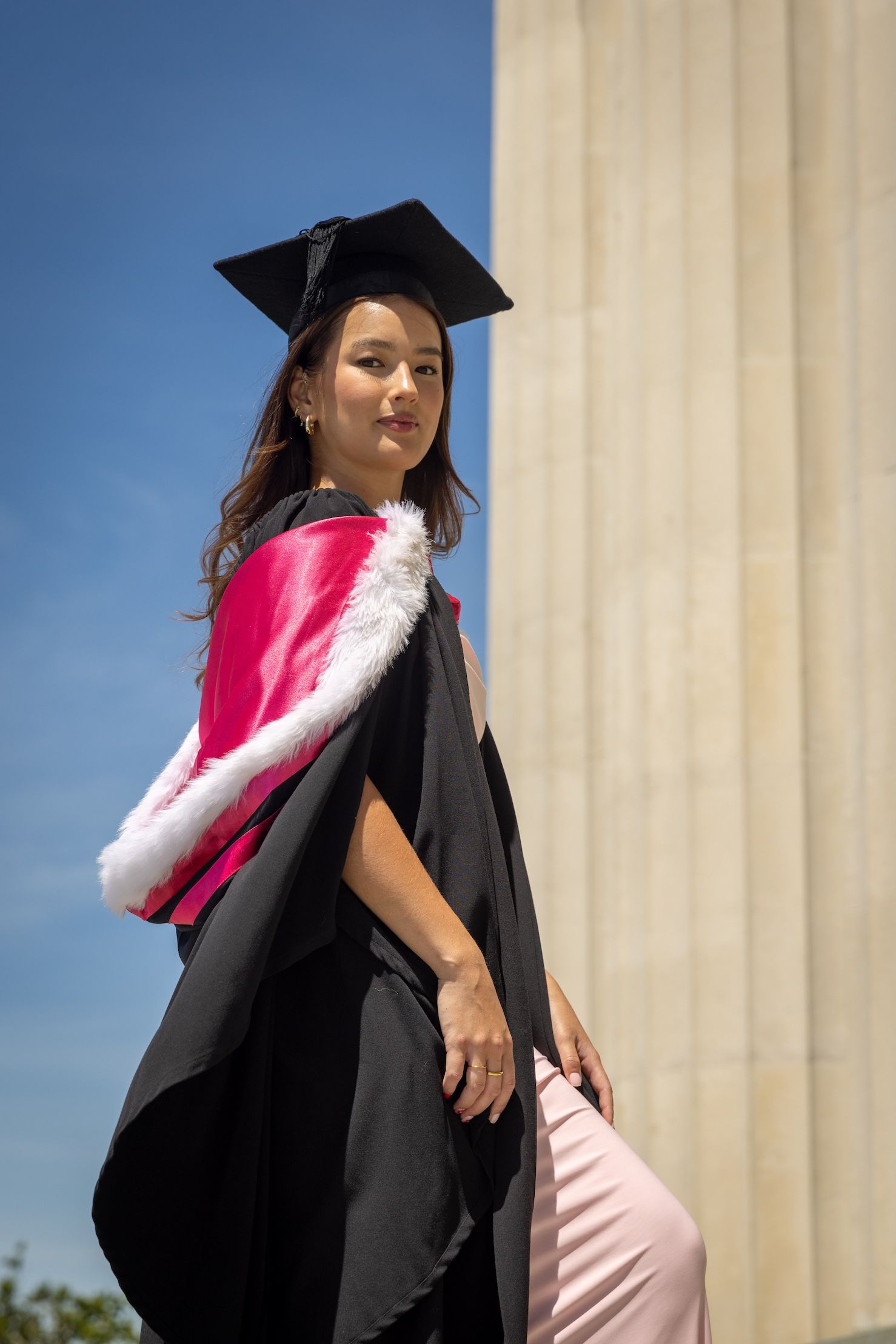 Young woman in black graduation gown and cap with pink hood poses proudly outdoors on a sunny day.