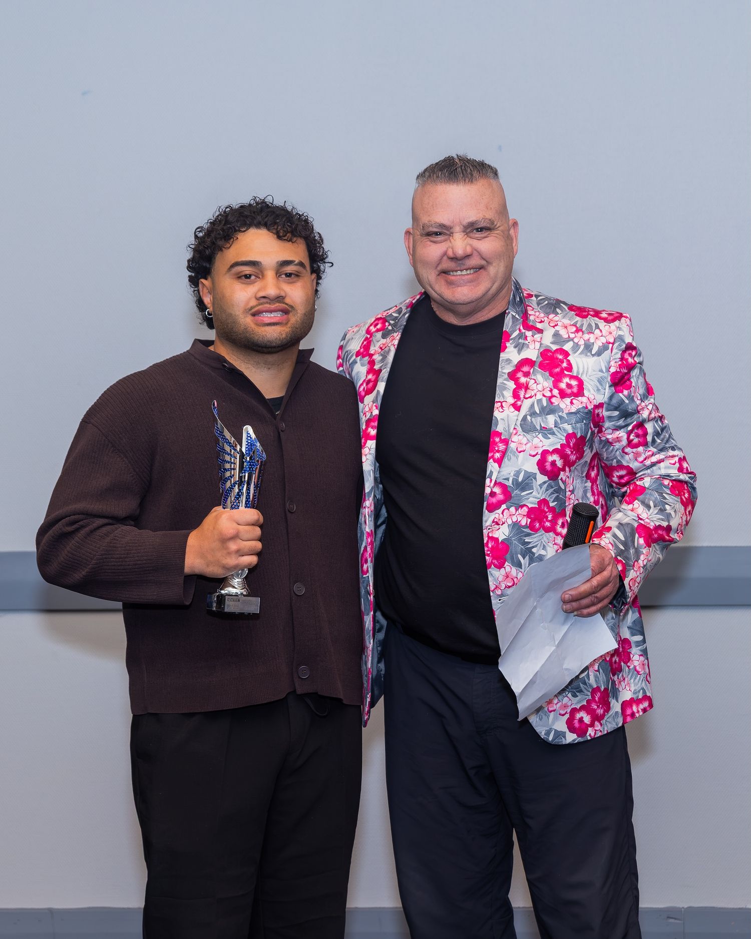 Two men pose together at an awards ceremony, one holding a trophy, wearing a floral blazer and dark outfit.