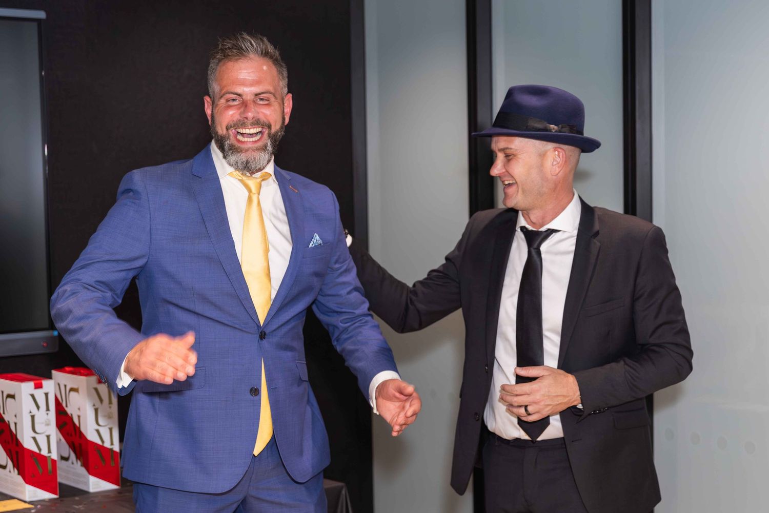 Two men in suits smiling and shaking hands, one in a blue suit with gold tie, the other in black with a hat.