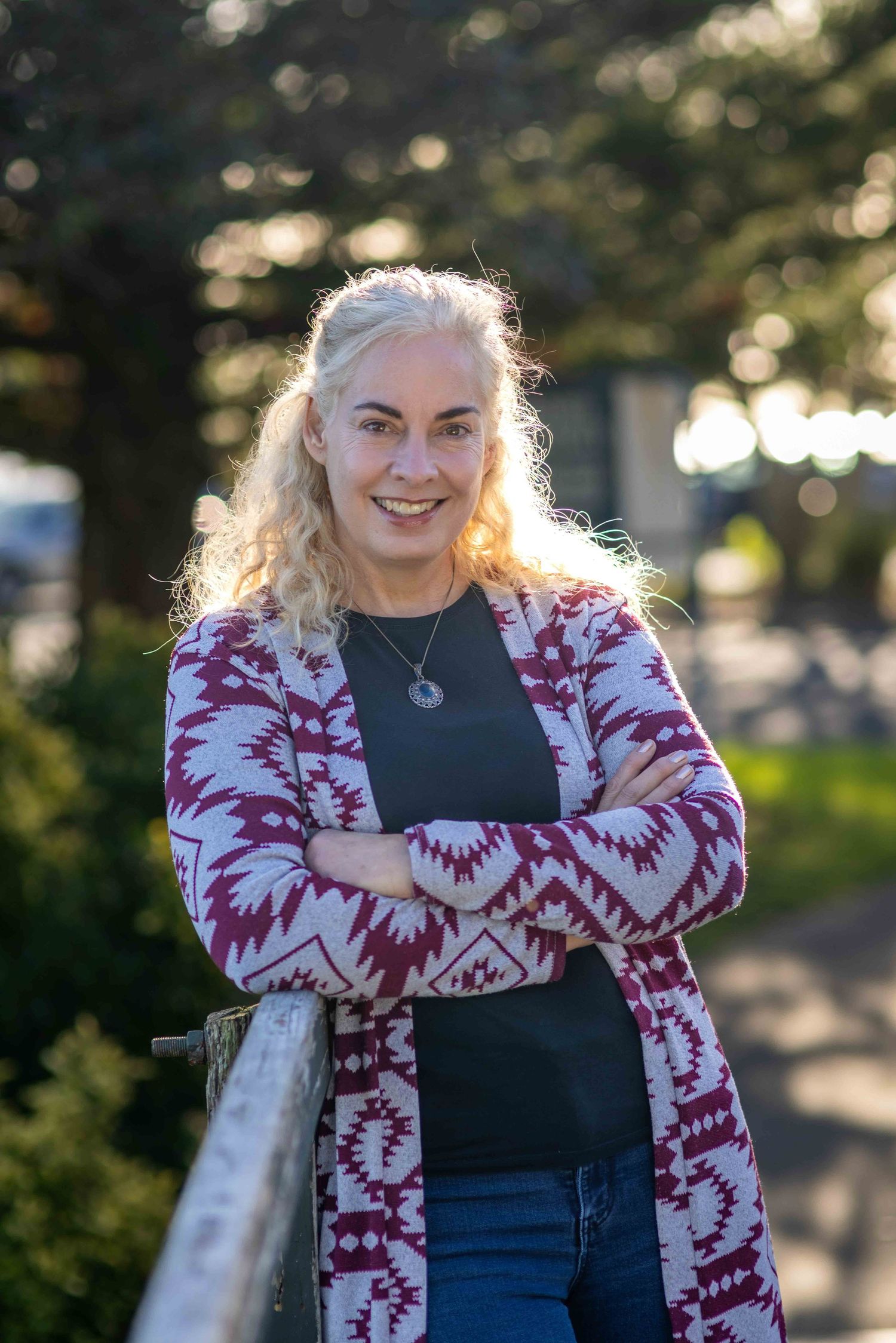 Smiling blonde woman with crossed arms wearing a patterned cardigan, posing outdoors in natural sunlight.