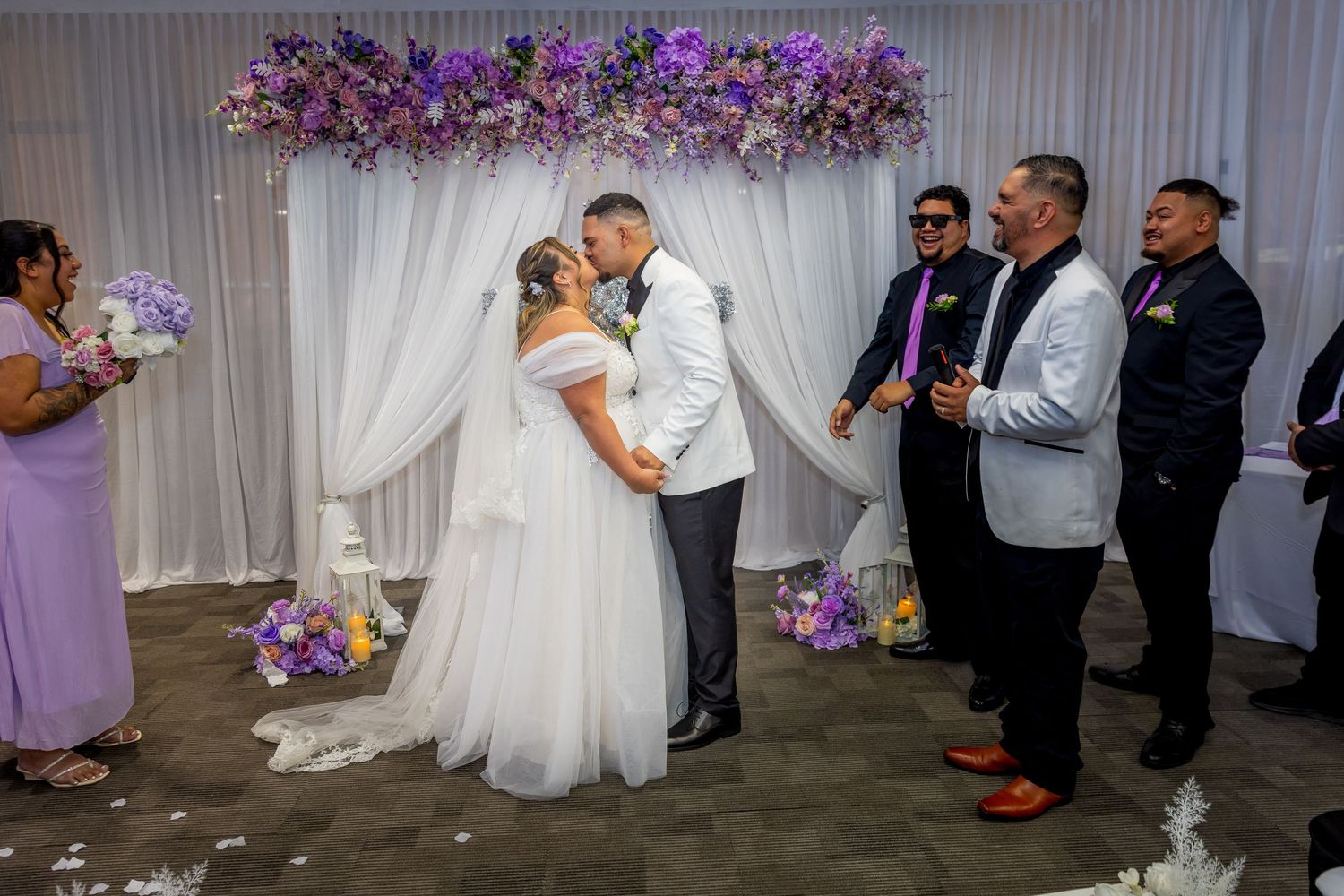 Bride and groom share first kiss at wedding altar adorned with purple floral arch, surrounded by guests in formal attire.