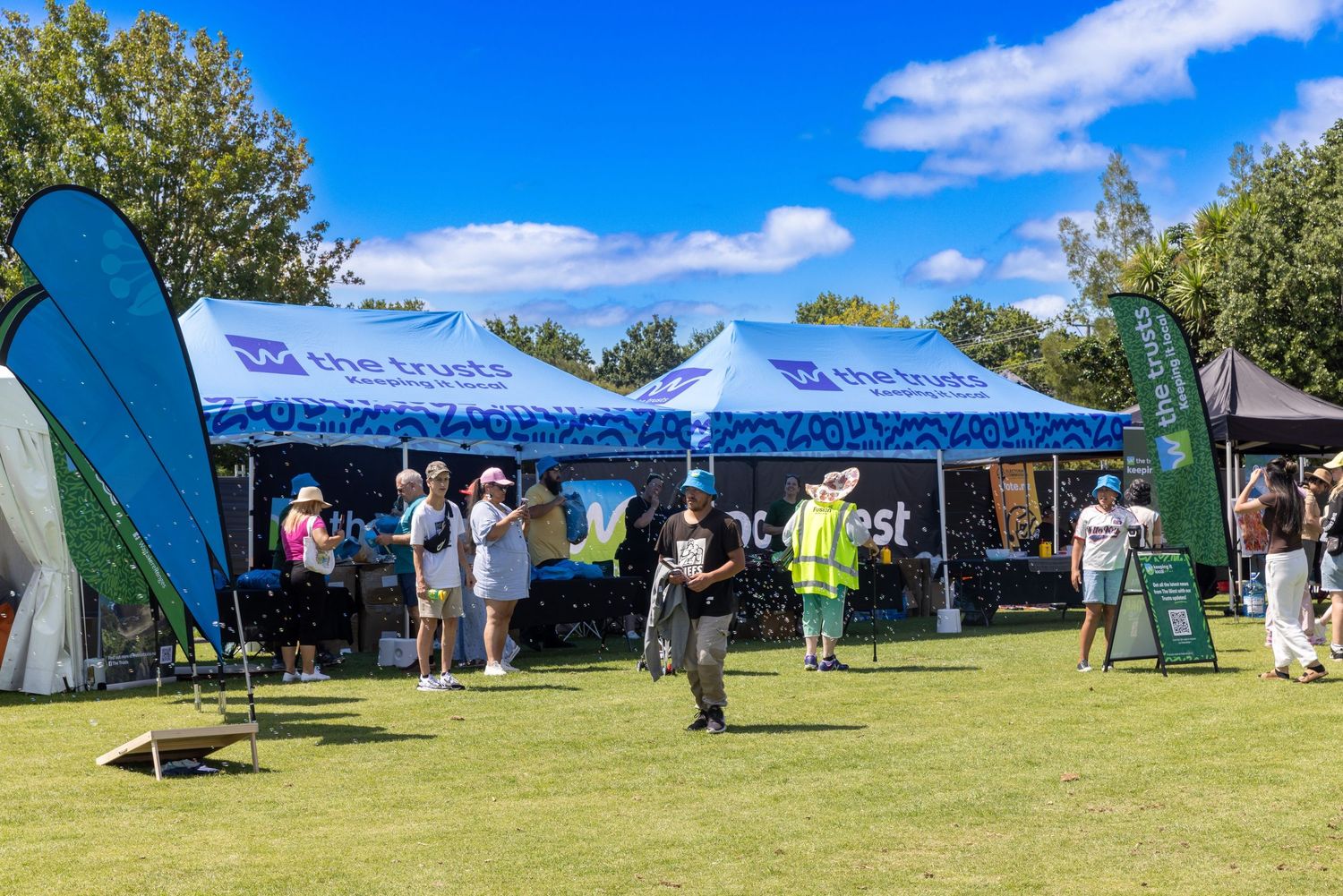 Blue and white event tents with promotional flags and visitors on sunny grass field.