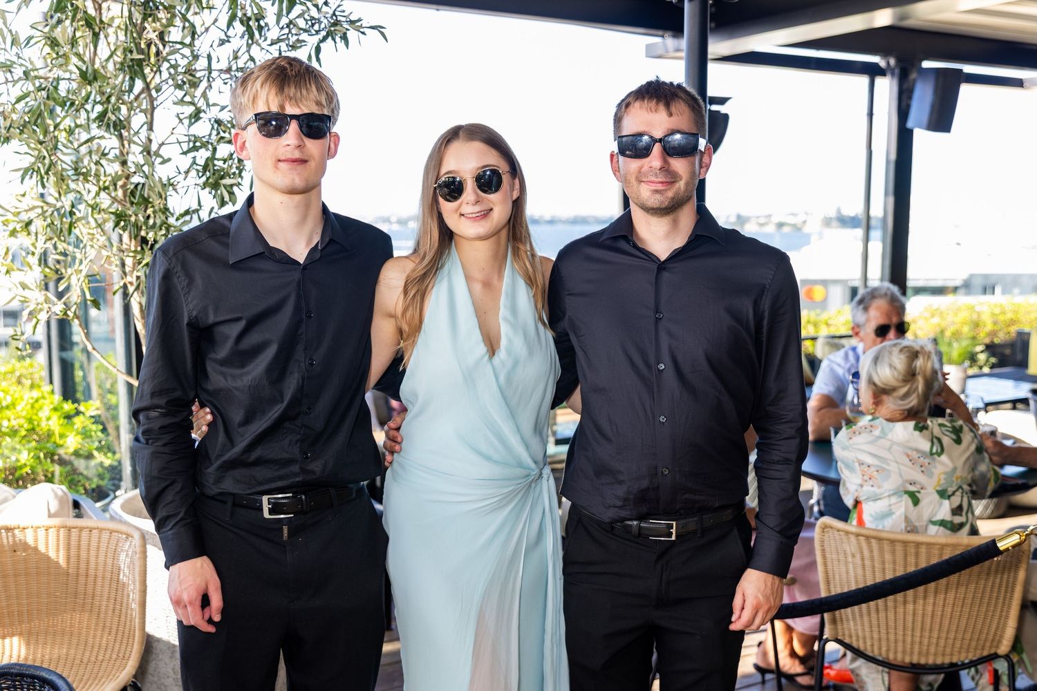 Three friends posing outdoors at a rooftop venue, two men in black shirts and a woman in a light blue dress.