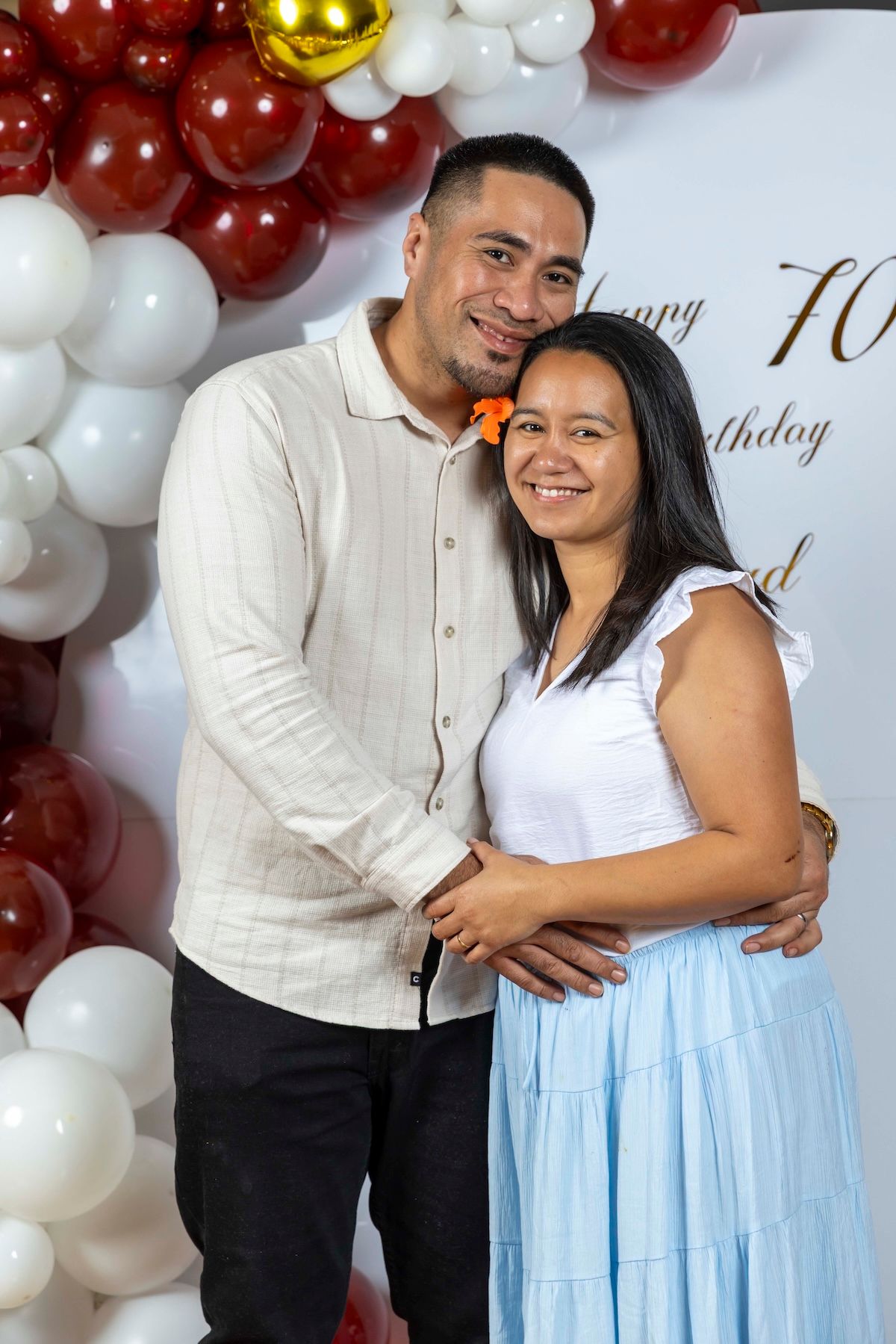 A couple poses together by a festive balloon arch with red, gold and white balloons at a 70th birthday celebration.