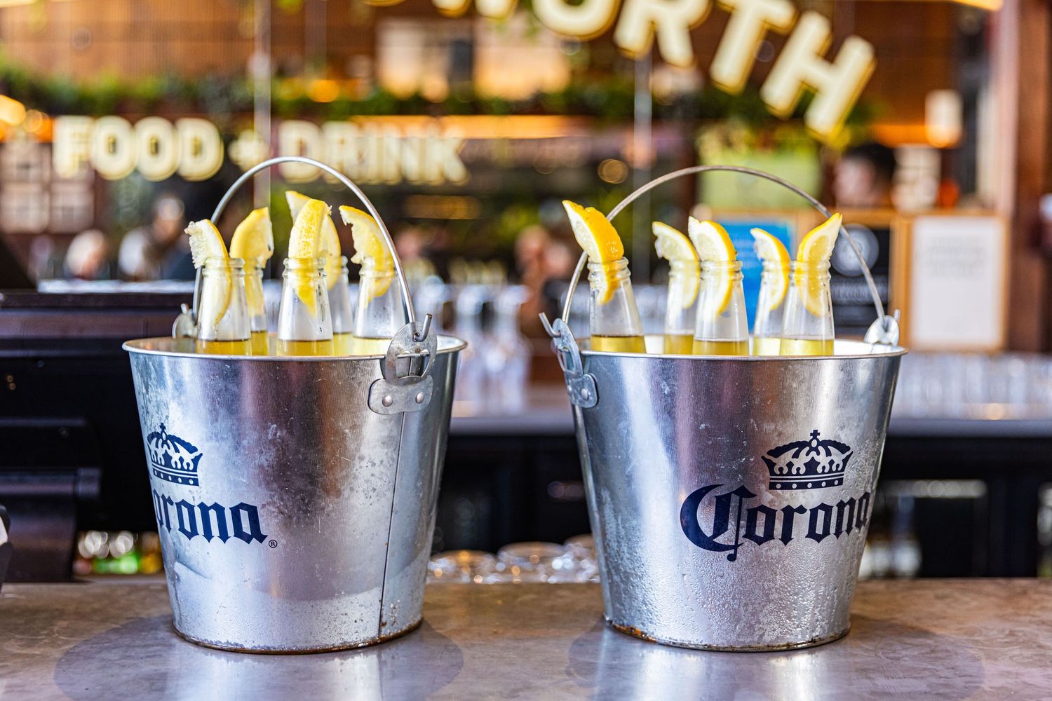 Ice cold Corona beers in metal buckets lined up on a wooden bar counter in a restaurant setting.