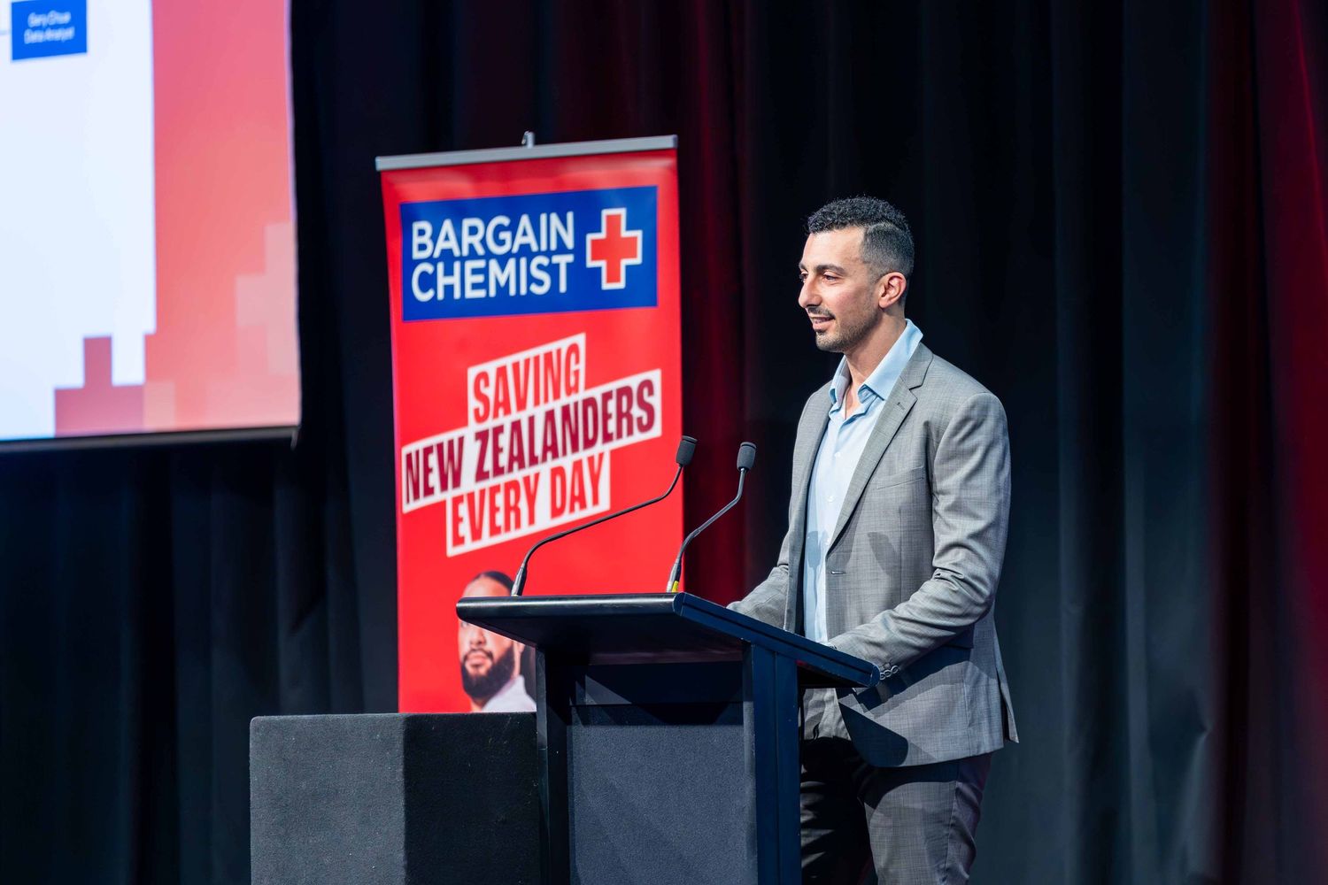 Professional speaker presenting at a Bargain Chemist corporate event with red branded signage in background.