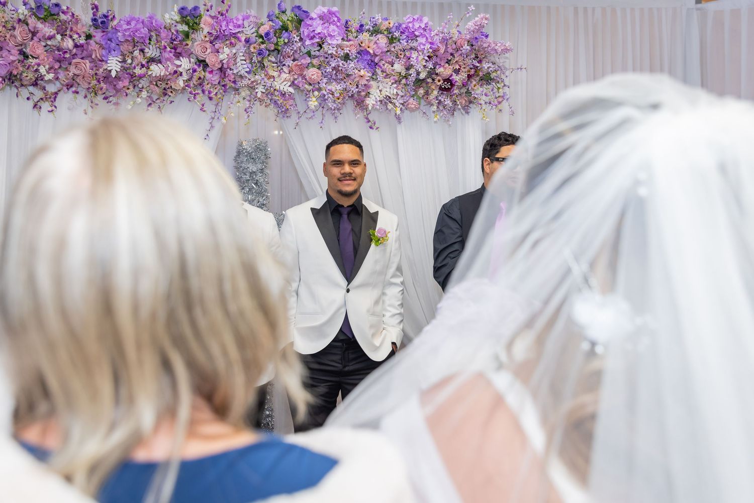 Groom smiling at bride walking down the aisle at elegant wedding ceremony with purple floral arch.
