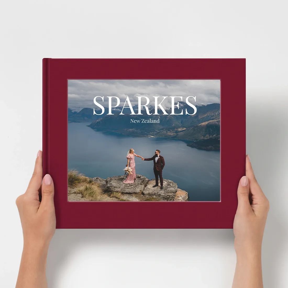 Hands holding a burgundy hardcover photo book titled Sparkes New Zealand featuring a couple on a cliff overlooking a scenic lake.