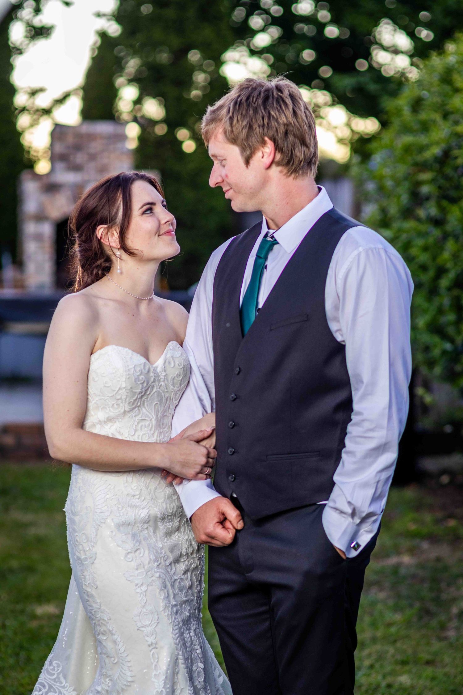 Bride in lace gown and groom in black vest with teal tie share a romantic outdoor moment at their wedding.