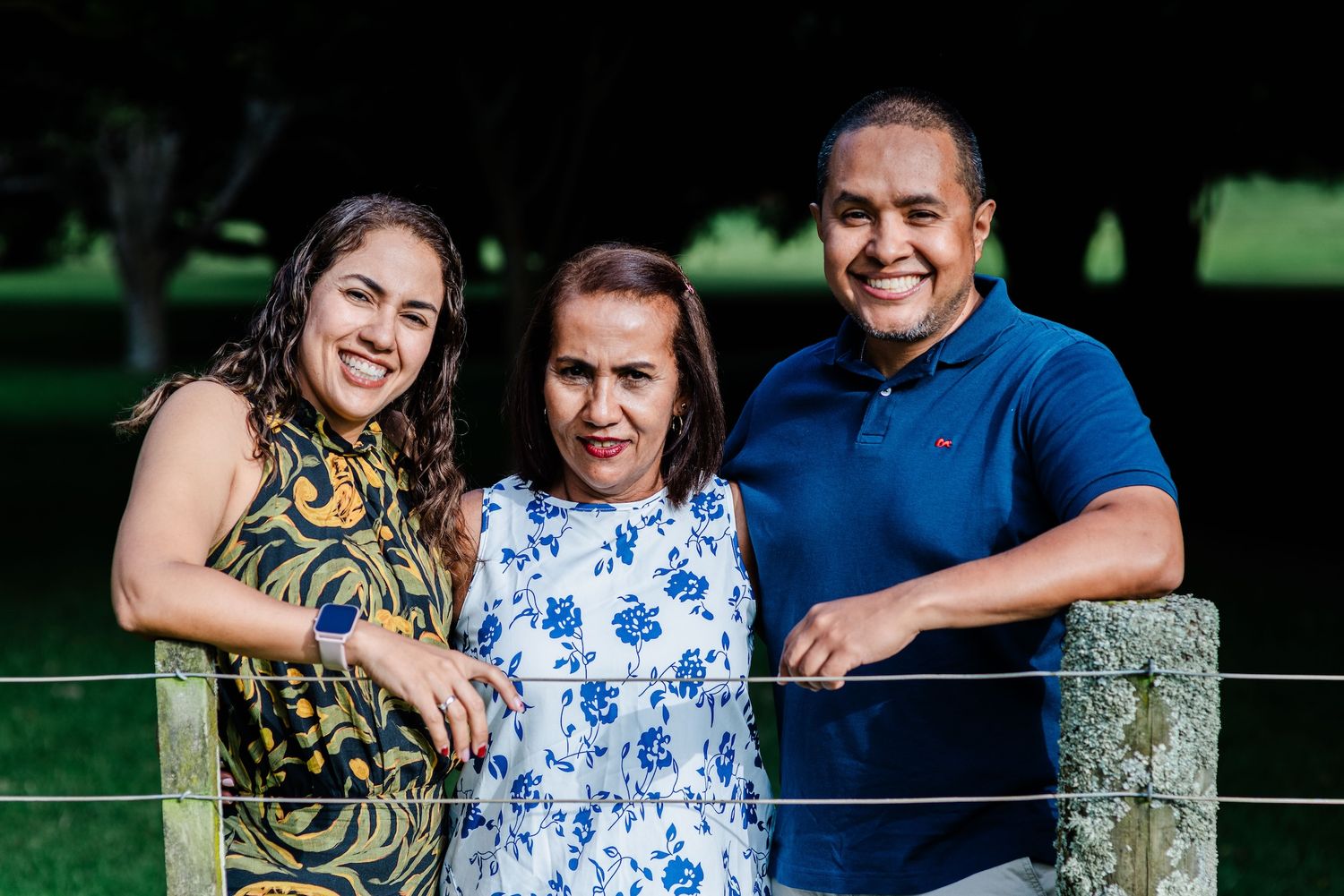 Three smiling people posing together outdoors by a wooden fence with lush green trees in the background.